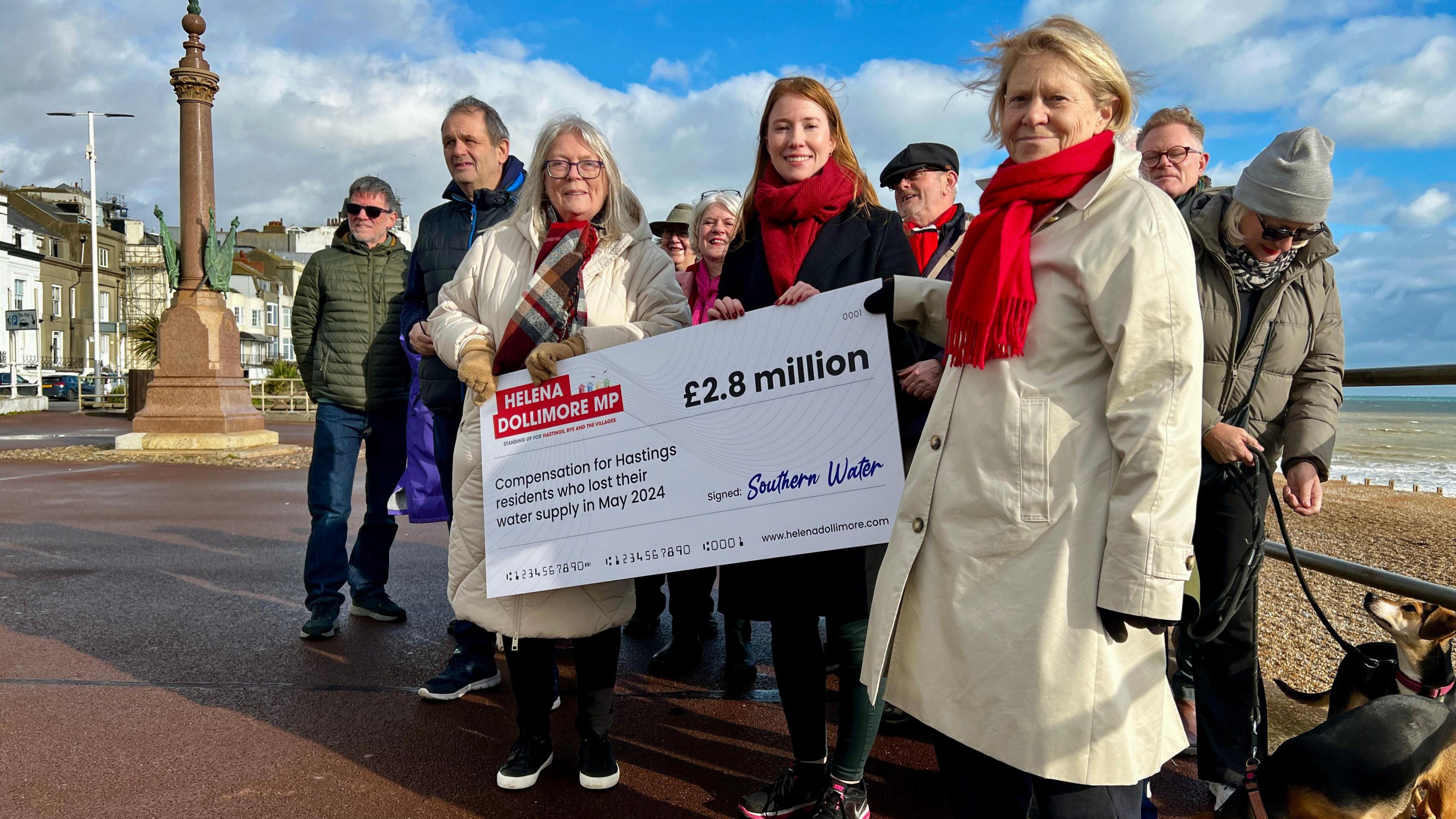 A group of residents stand around a large cheque for £2.8m signed by Southern Water and with Helena Dollimore's name at the top. They are standing on a  seafront with the beach visible in the background.