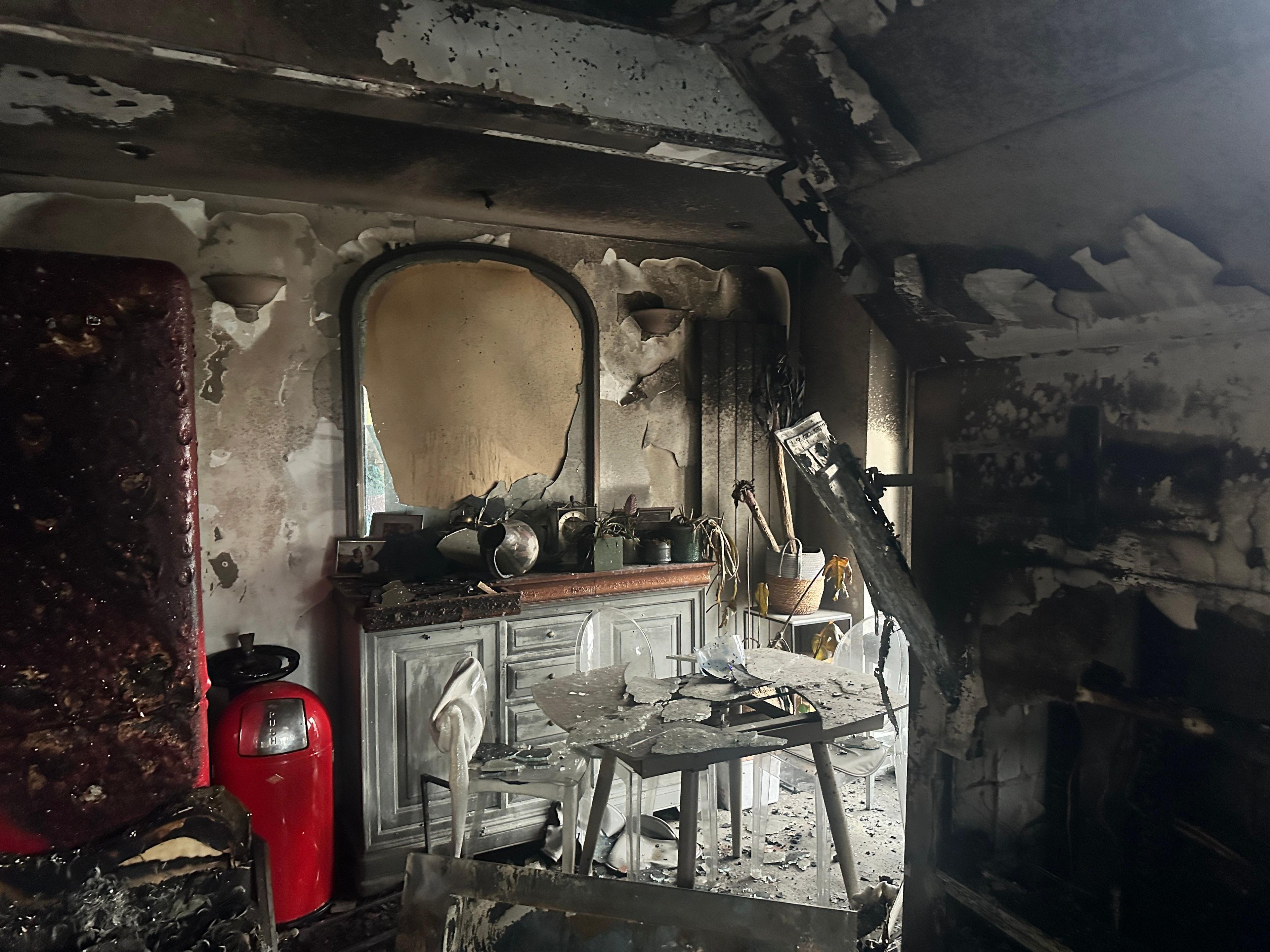 A burnt out kitchen. There is a red bin in the left-hand corner of the room. The is wallpaper coming off and the red fridge is a melted plastic on the left side of the room. In the middle there is a table and chairs with charred remains of the room.