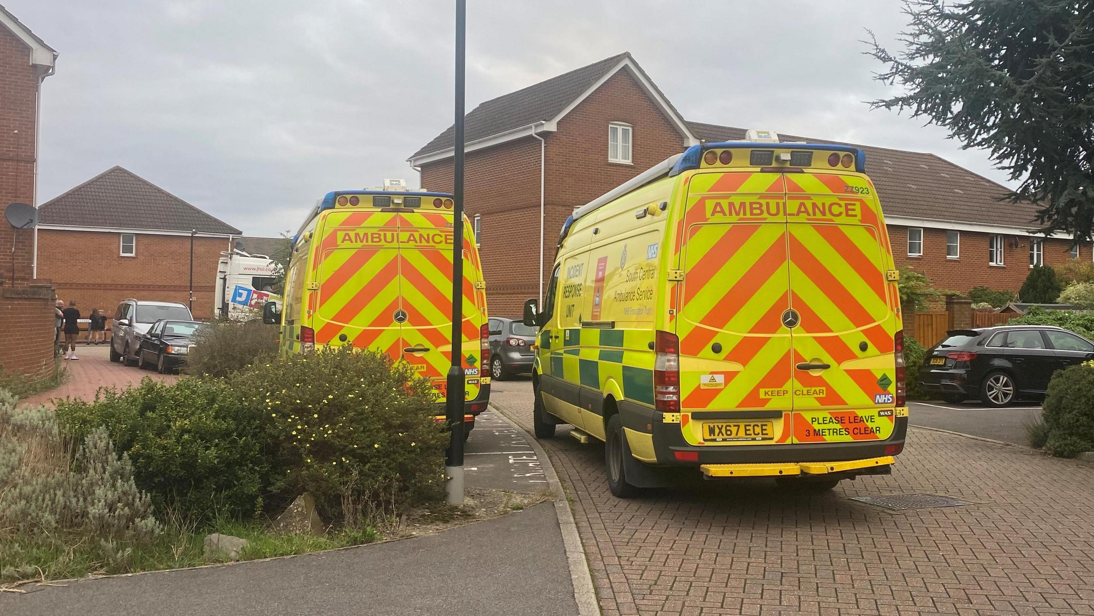 Two yellow ambulances parked on the side of the road by some bushes. Around them are houses, with several people stood at the side of the road further down