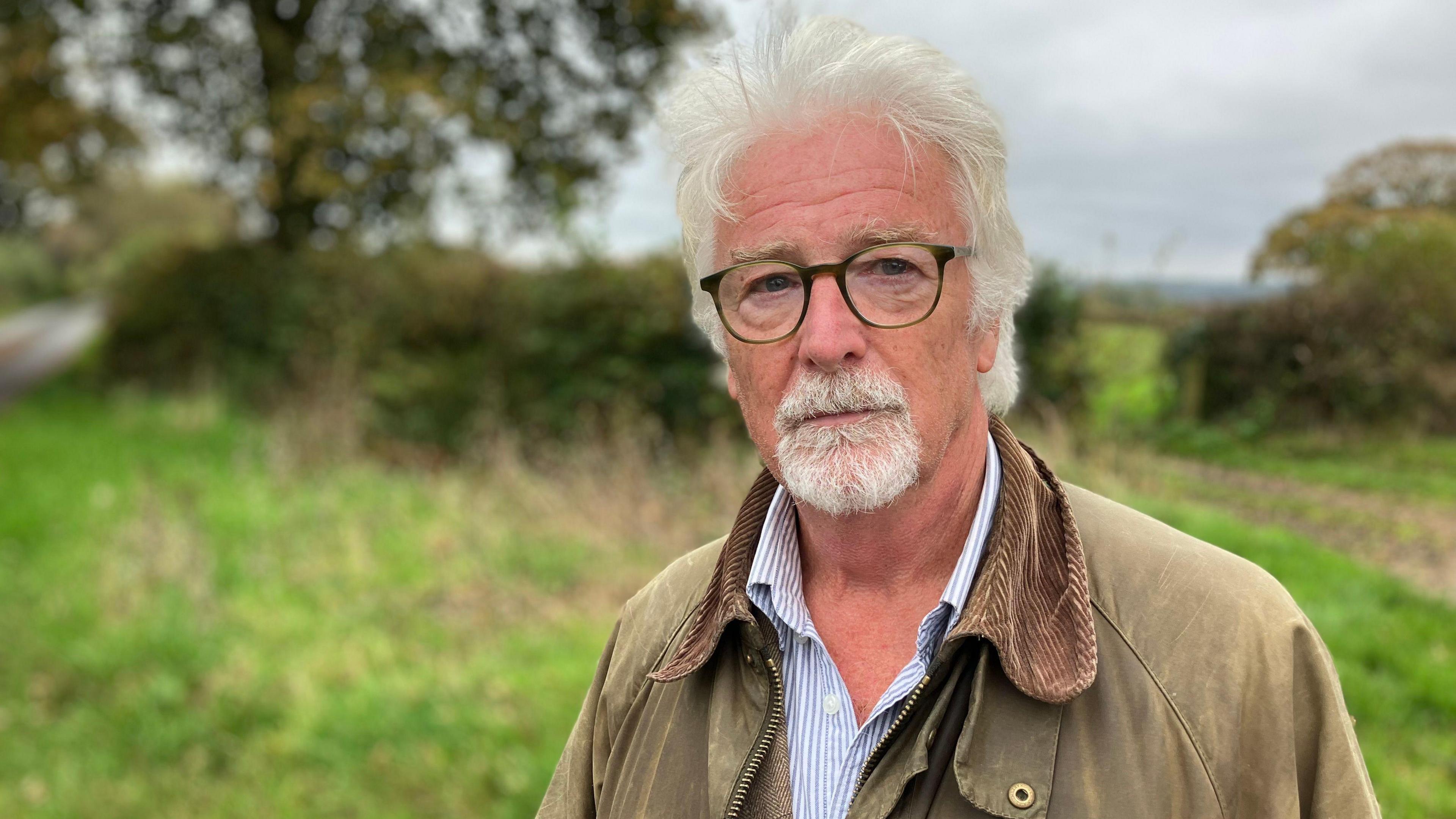 Stephen Crowther stands in a country lane. He has grey hair and grey beard and is wearing glasses. He is wearing a brown waxed jacket. Behind him is the entrance into a field and a green roadside verge.