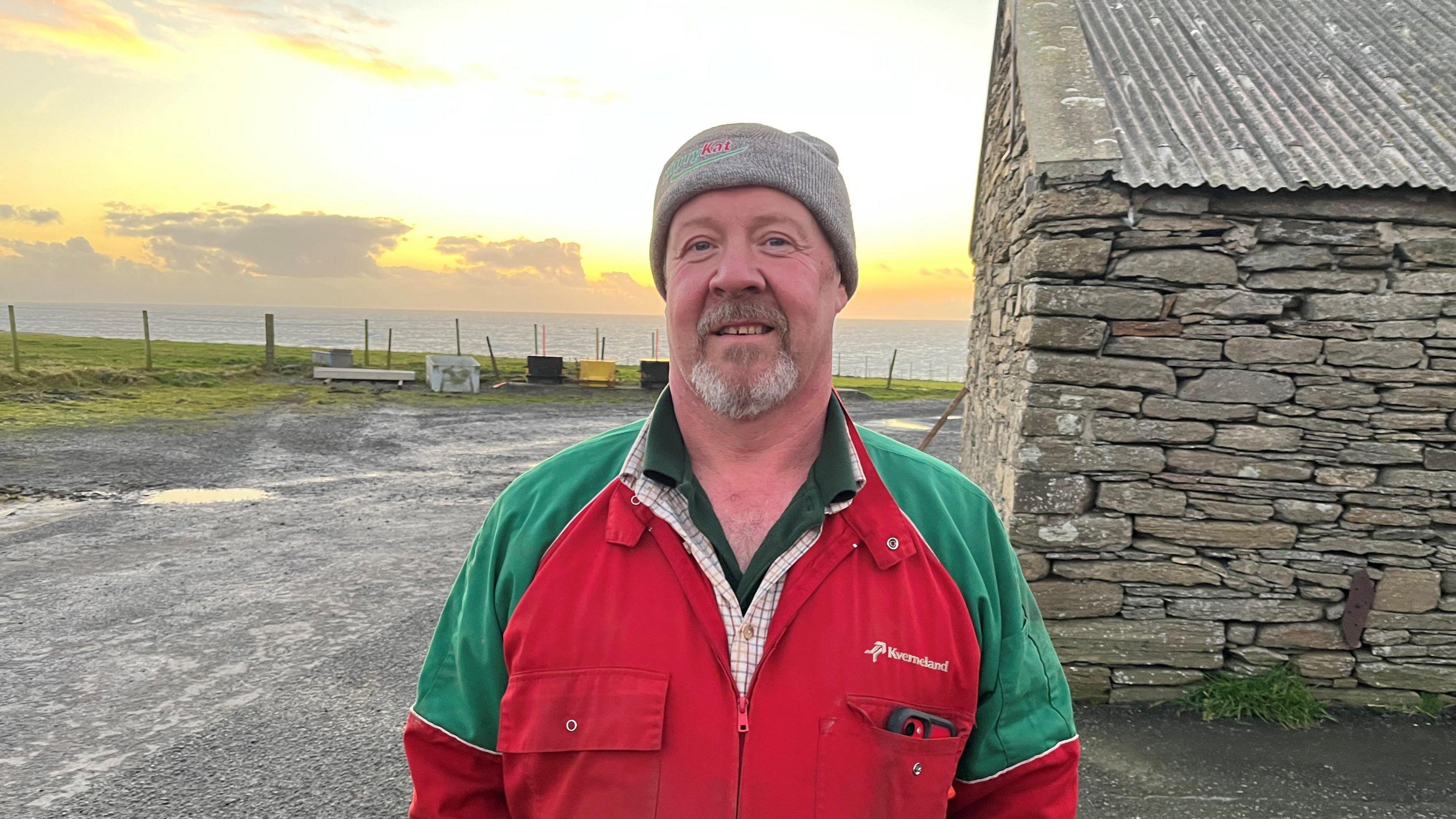 Man smiling at the camera with a stone building and the sea and a yellow sky behind him. He has a beard and is wearing red and green overalls over his shirt and a grey woolly hat. 