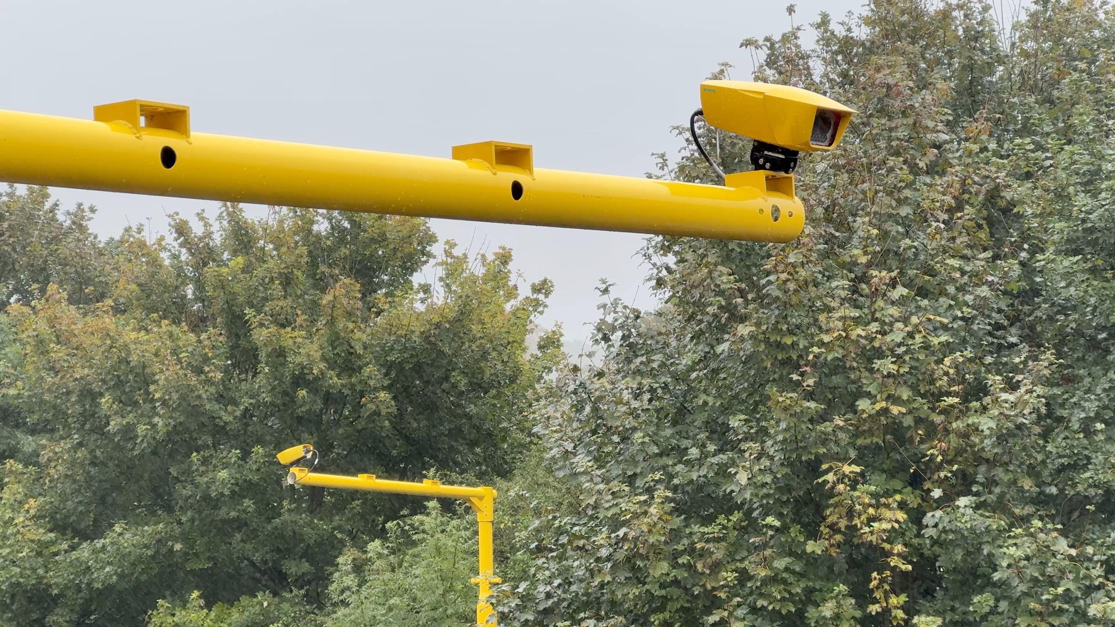 A yellow average speed camera. The camera extends over the road from a yellow pole. The camera is seen facing down towards the road.