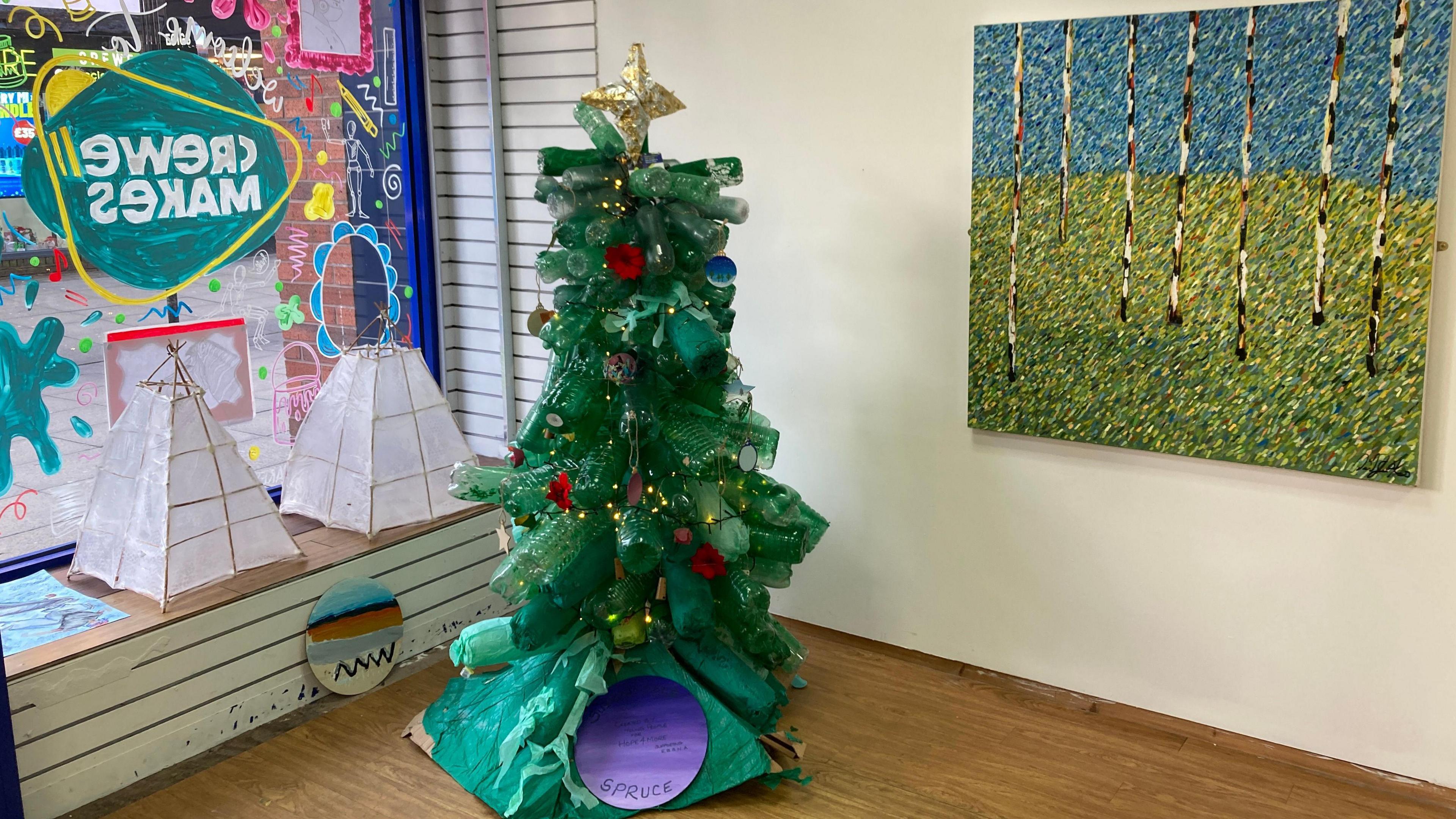 A sustainable Christmas tree made from empty plastic bottles, in the concert of Crewe Makes, with a window display on the left and a painting on a wall to the right.