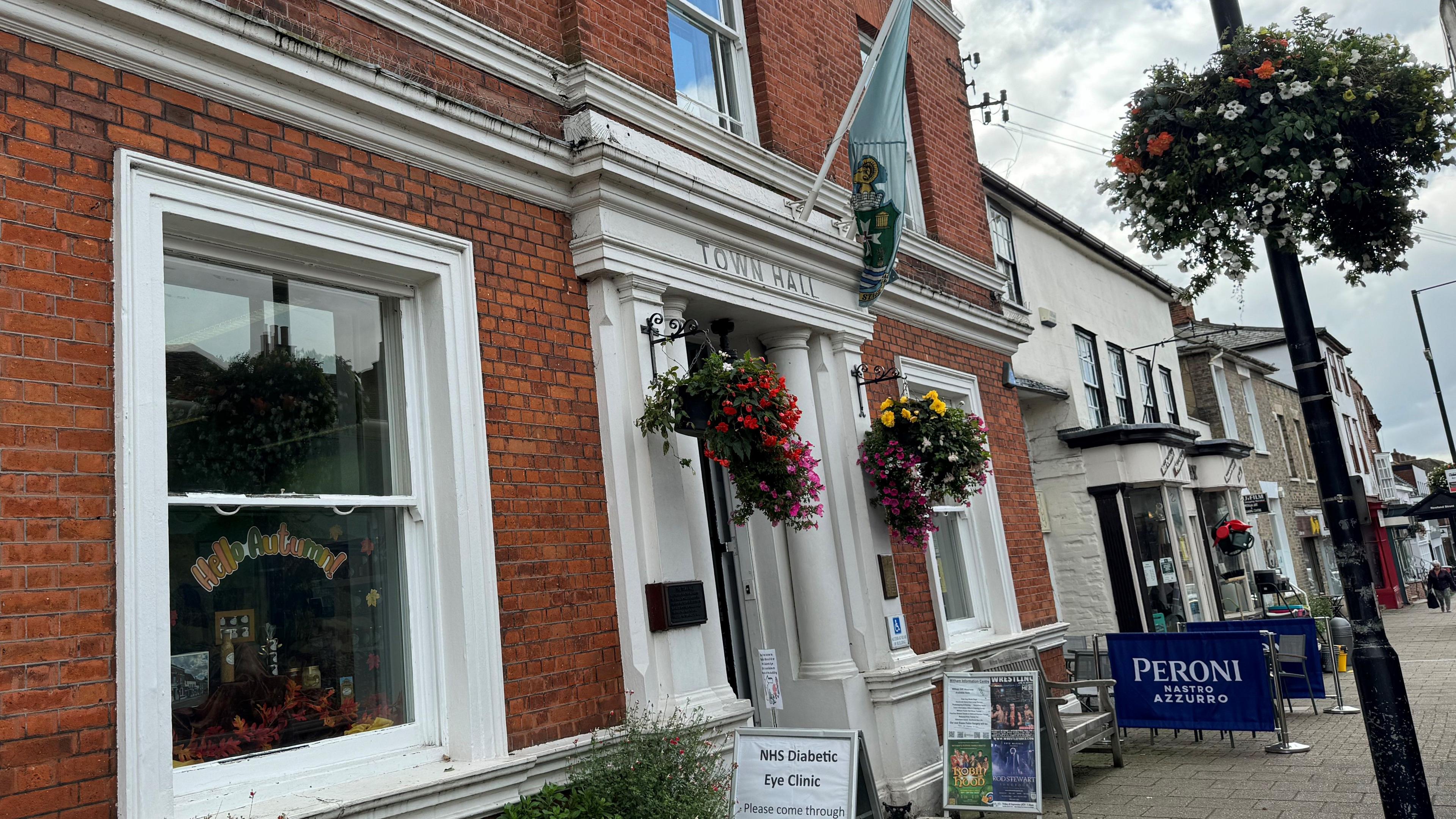 The front of Witham Town Hall, where there are hanging baskets with flowers at the front door and a flag above the entrance.