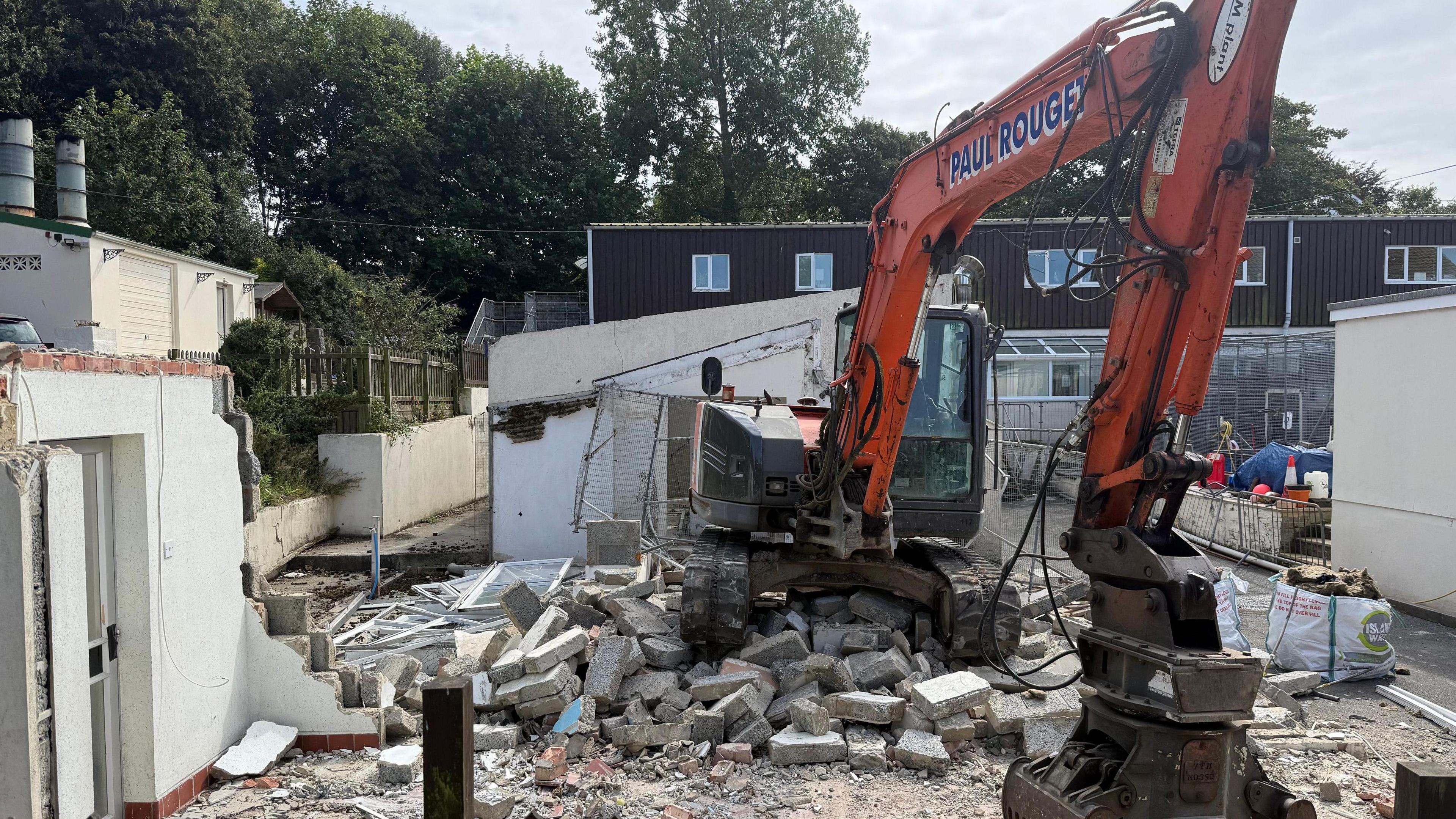 A small digger is parked on a large pile of pieces of concrete with its bucket resting on the ground. A building to the left has been partly dismantled. Behind the digger is a two-storey building.