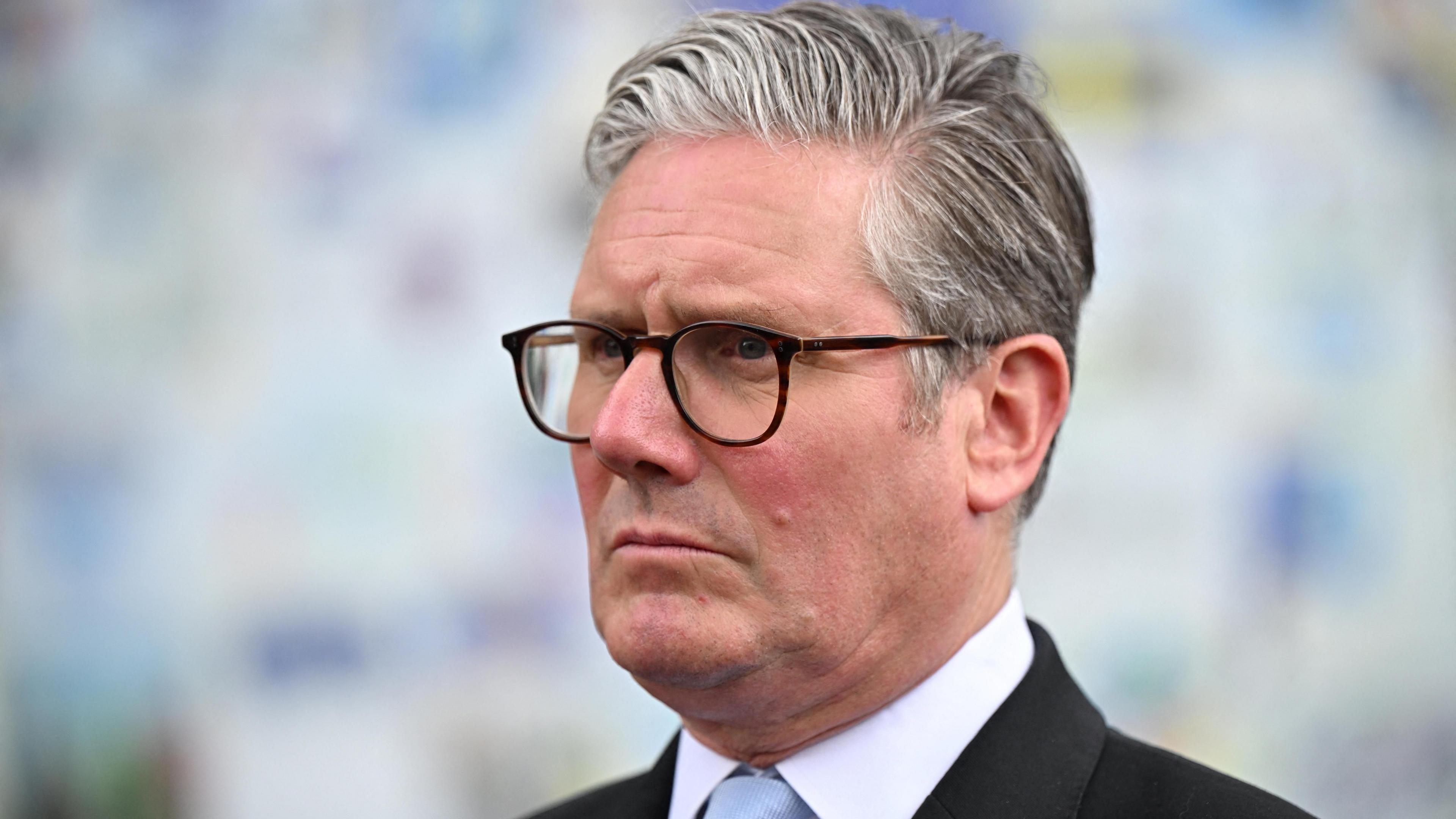 A close up of Sir Keir Starmer, wearing glasses and a slightly perturbed expression. A pale blue tie can be glimpsed beneath a white collar and dark blazer. 