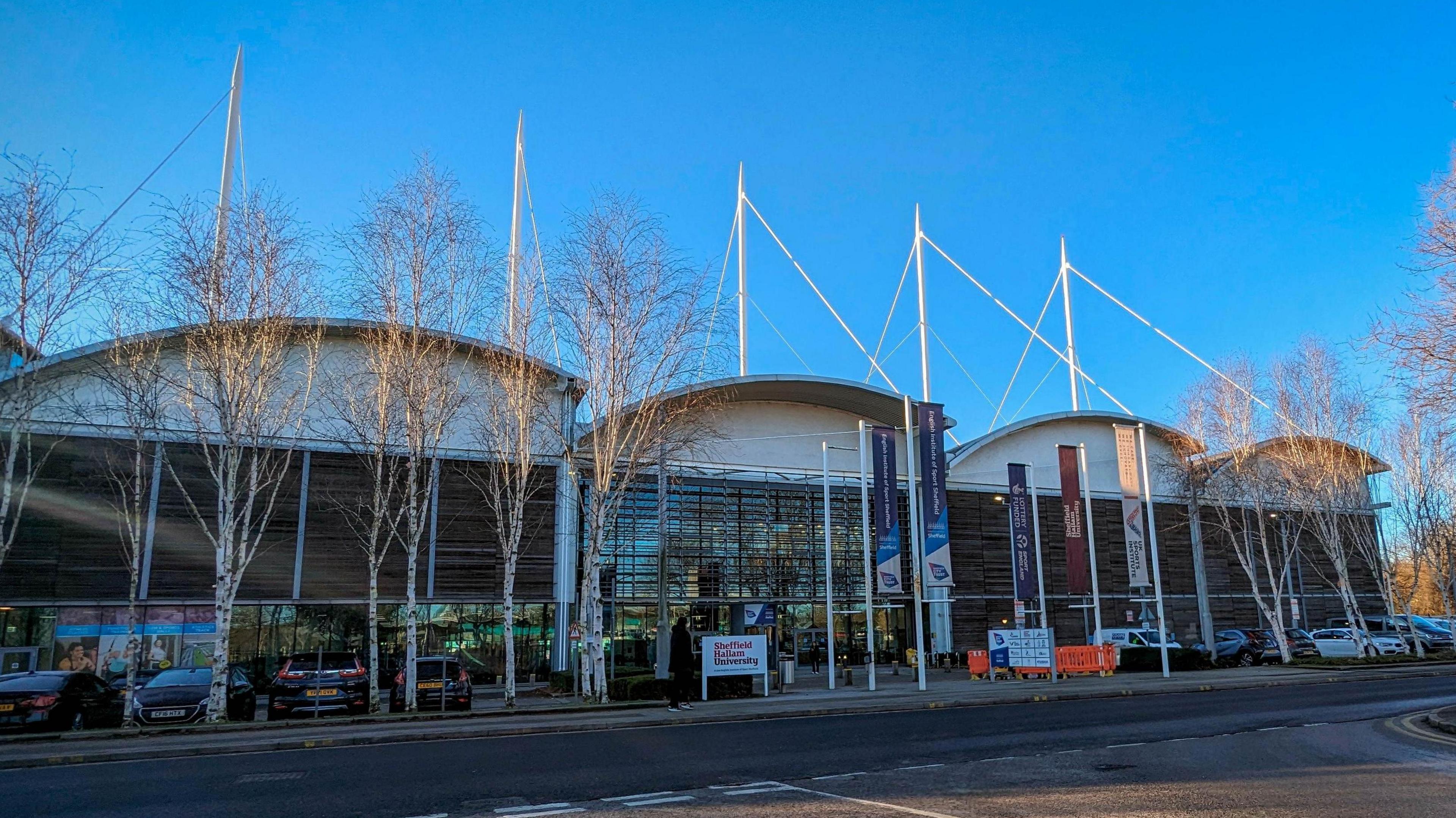 The exterior of a large modern sports facility with sloping arched roofs and glass panels