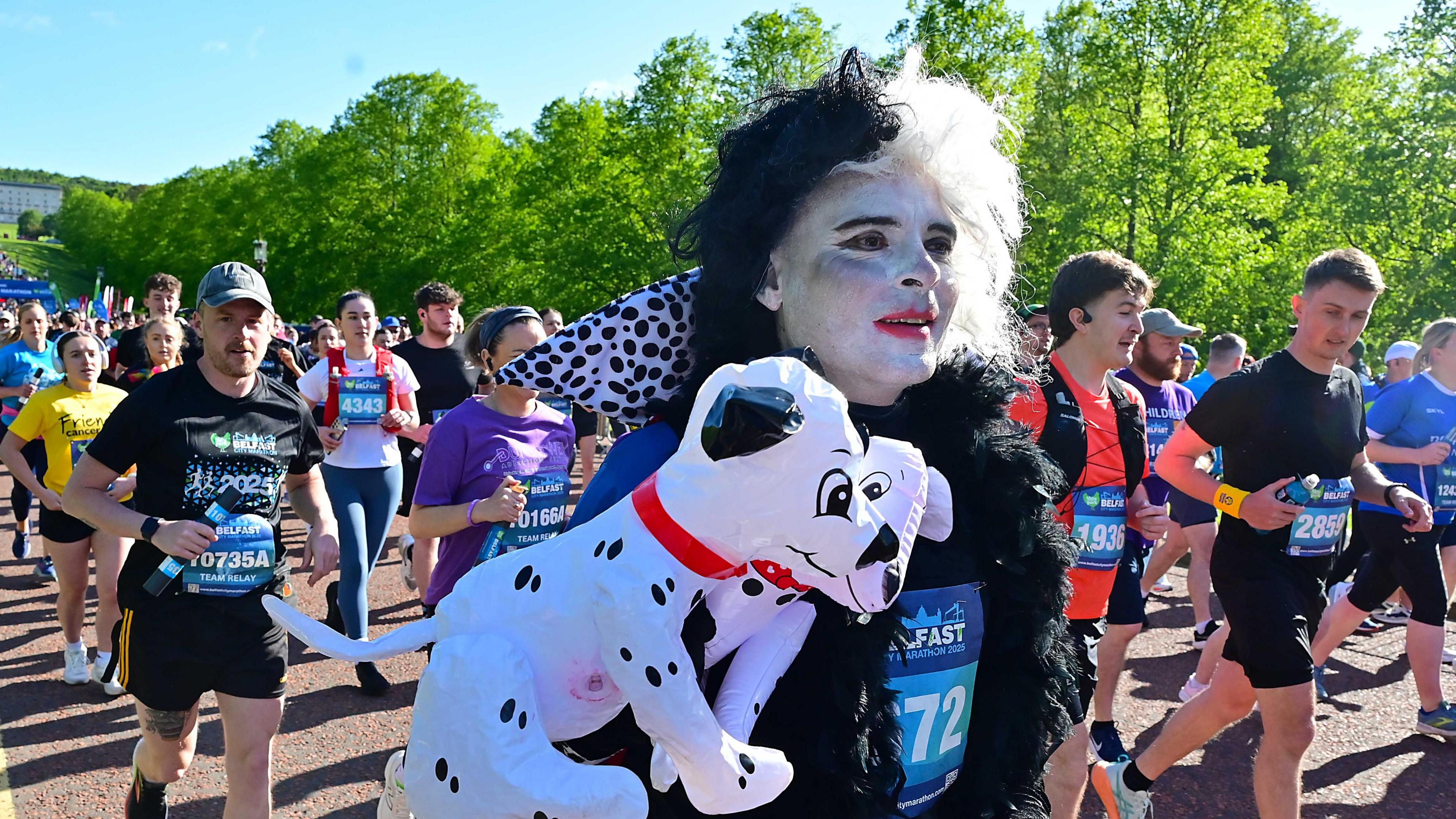 At the start line of the marathon is someone dressed up as Cruella de Vil. They also have inflatable black and white Dalmatians. Behind them is numerous runners beginning the marathon.