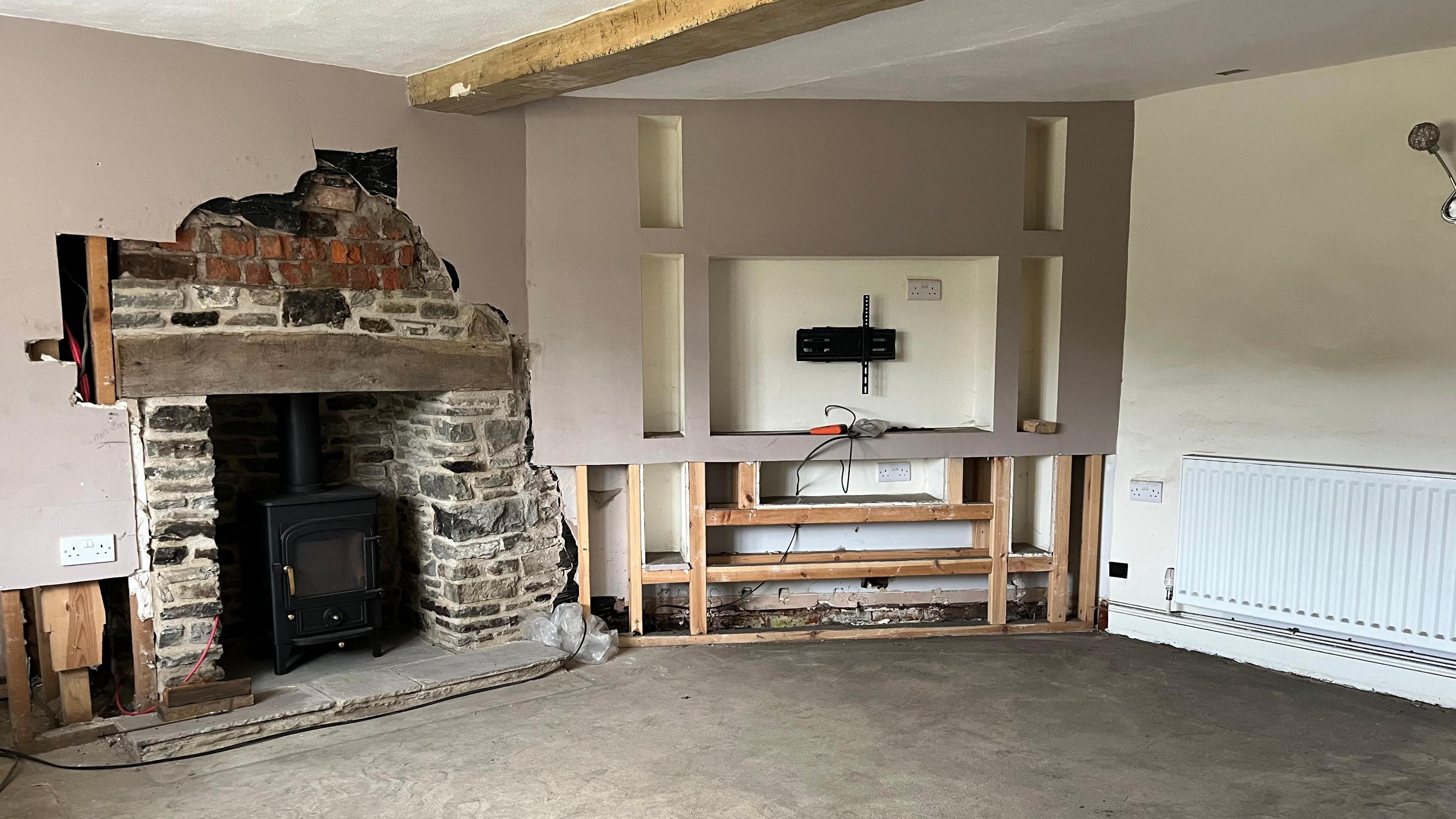 Photograph of what was Paul's living room. The far wall is beige and has patches of discolouration on it. The other two walls - one of which has been ripped out in the middle - are painted grey. The brick on the fireplace is exposed, and you can see cables trailing from where the TV has been taken off the wall. The floor is exposed, dusty concrete.