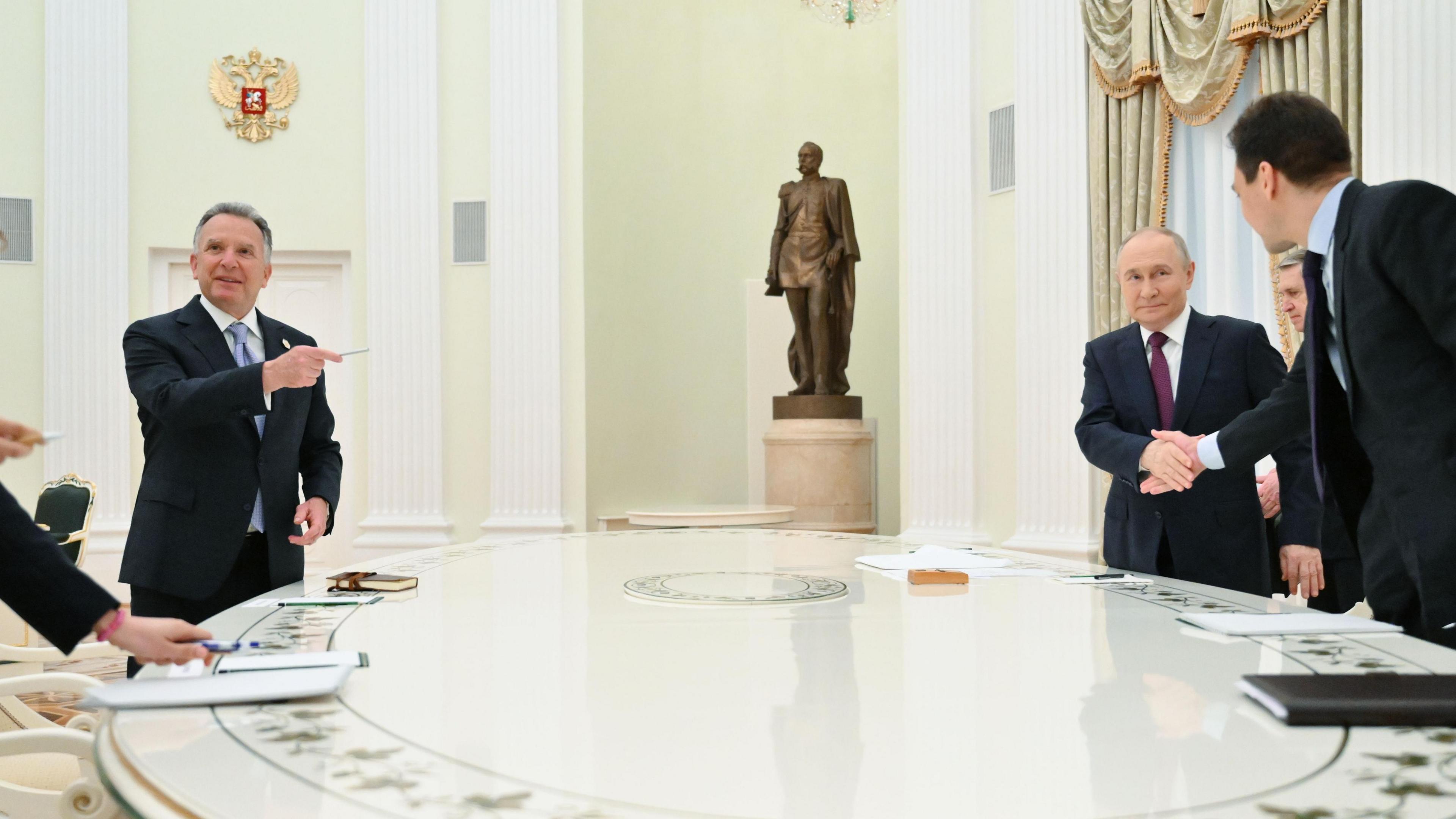 Witkoff, who wears a suit and has short grey hair, points with a pen towards Putin, who also wears a suit and had short thinning grey hair, as they stand around a large white table at the Kremlin