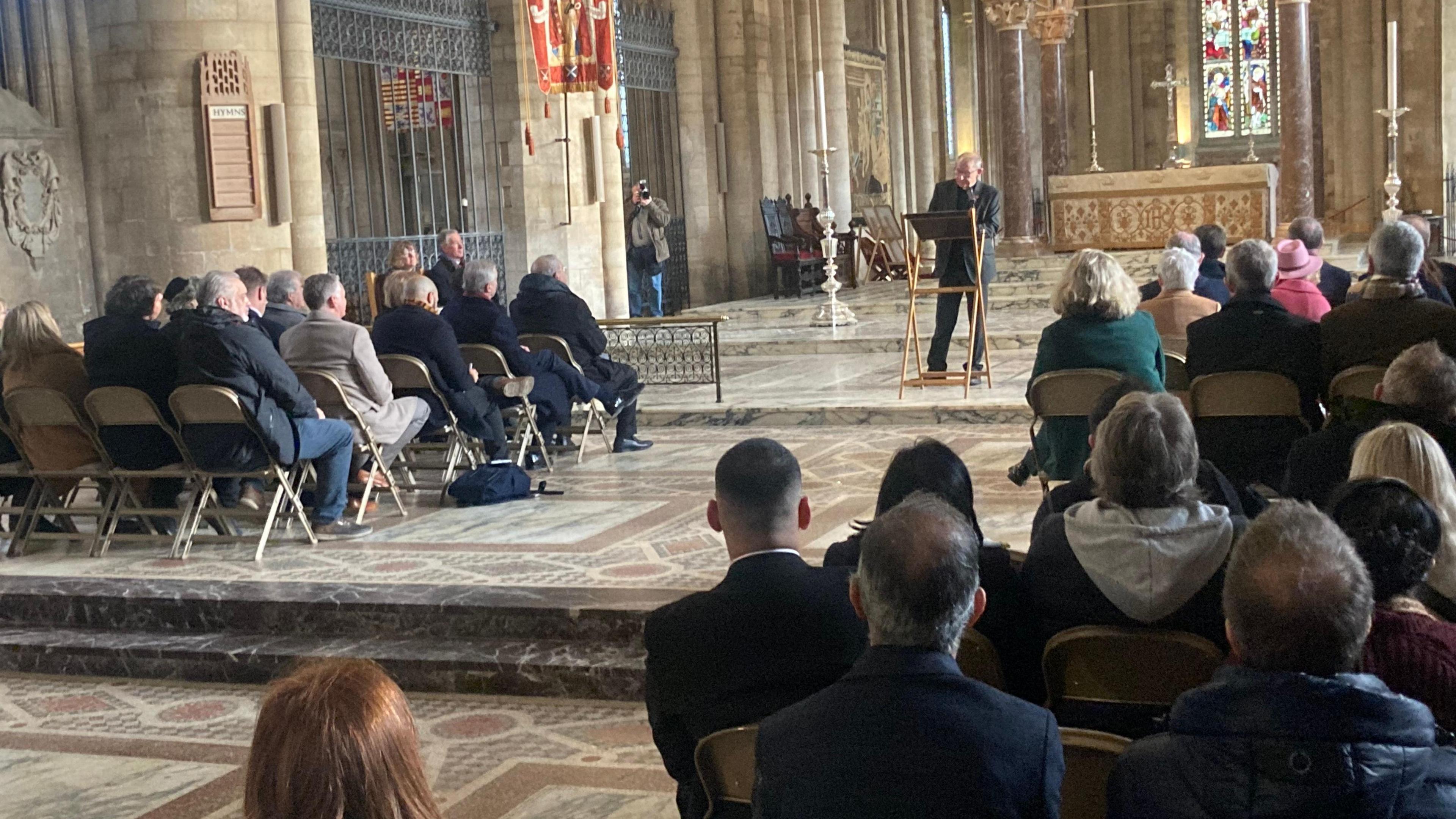 People on sitting on chairs on either side of the main room in the Cathedral with the Dean standing in the nave addressing the gathering 