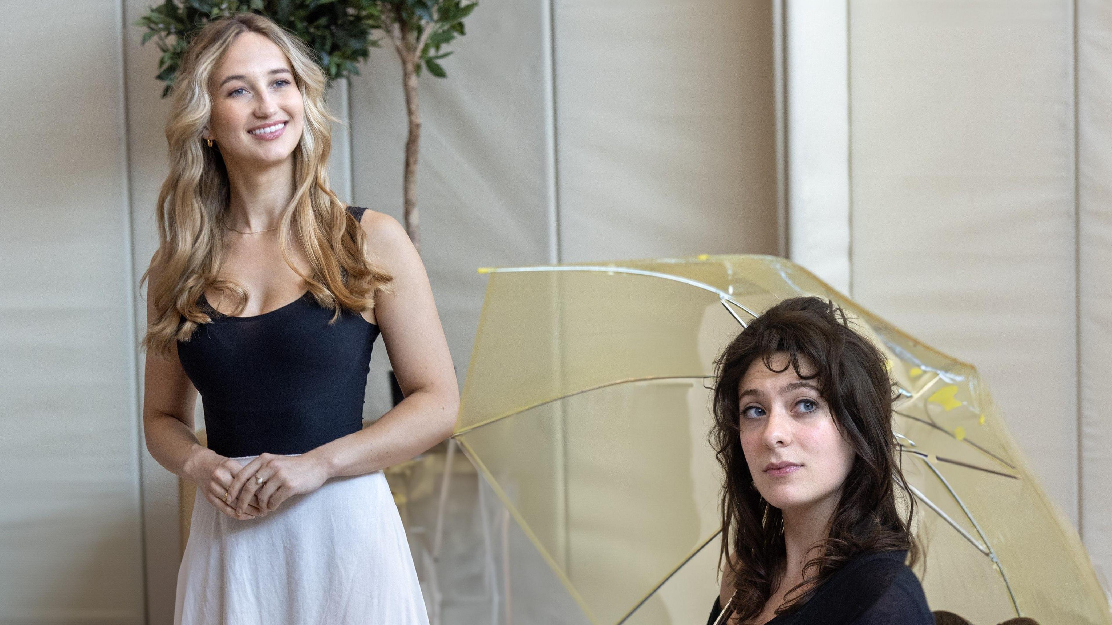 Two actresses, one with dark long hair, seated and holding a light yellow umbrella, and one standing with blonde hair and wearing a white skirt and dark blue vest top, take part in rehearsals for Emma at the Theatre Royal Bath