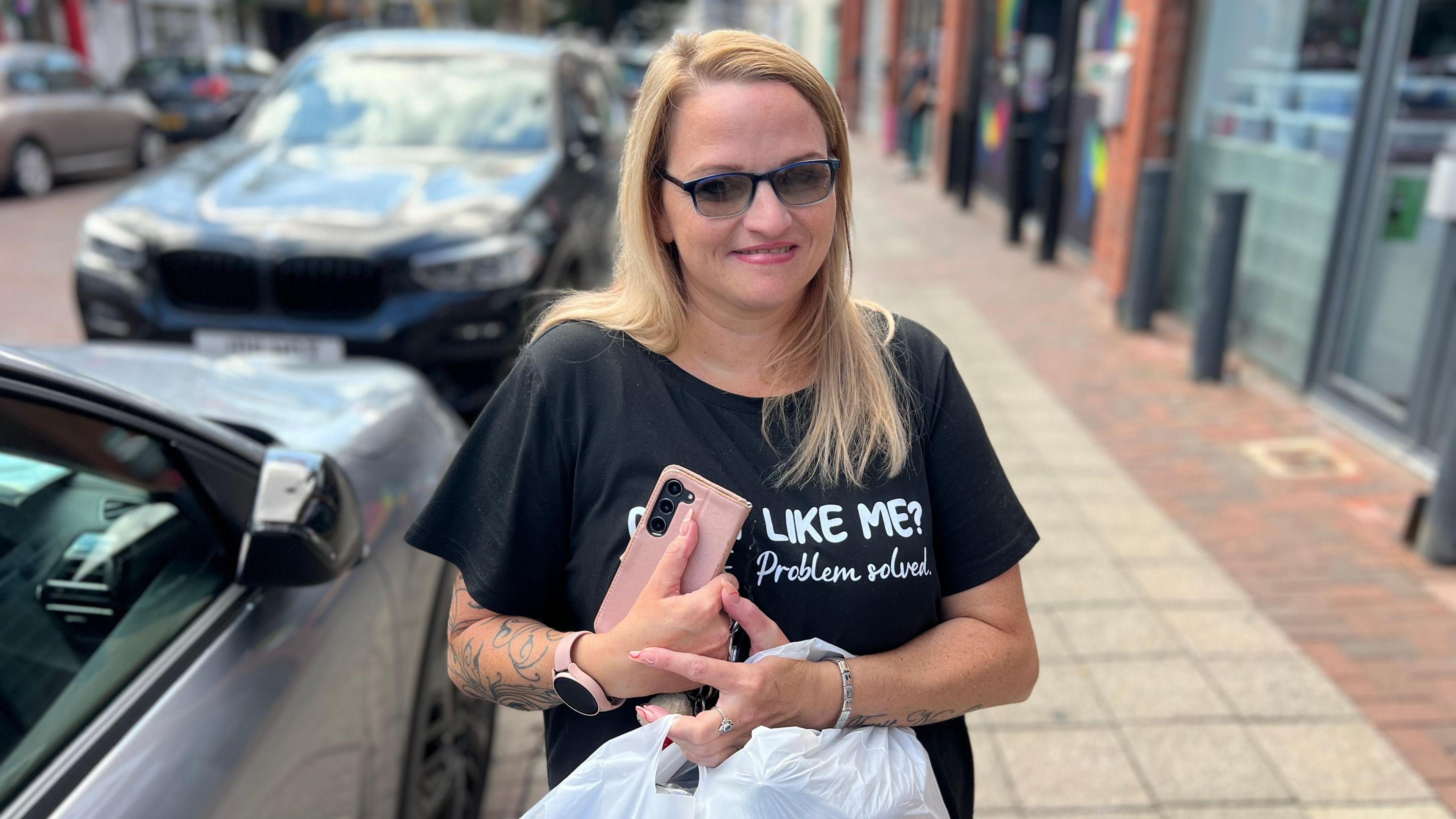 A woman with blonde hair and sunglasses holds her mobile phone and shopping bags as she smiles into the camera. She is standing on a street with shops on the right and parked cars on the left.