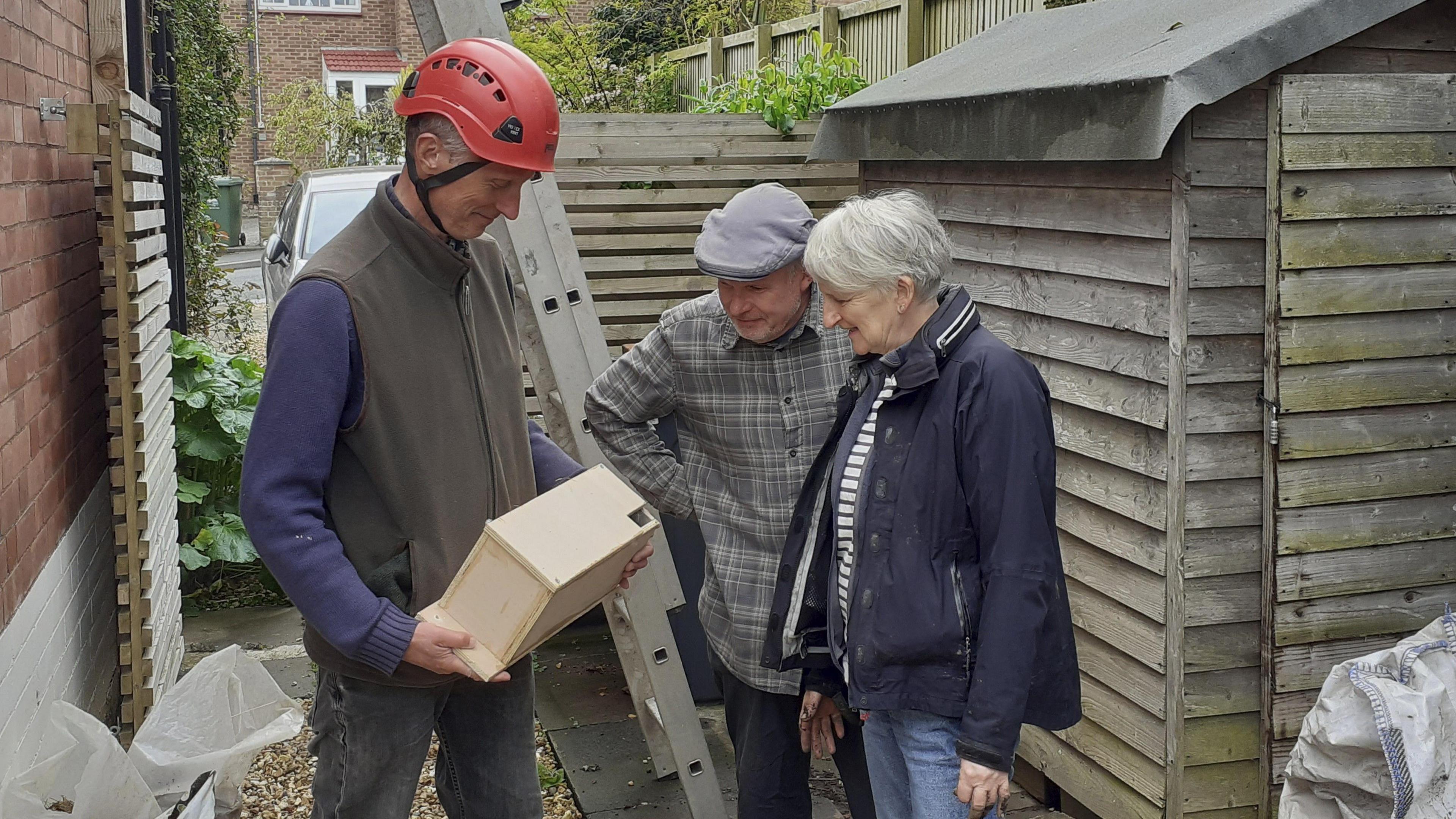 A man and a woman are standing next to another man, looking t a box he is holding. The box is wooden and made for swifts, with a small hole in the corner. He is wearing a red hard hat and there is a ladder leaning onto the side of a house