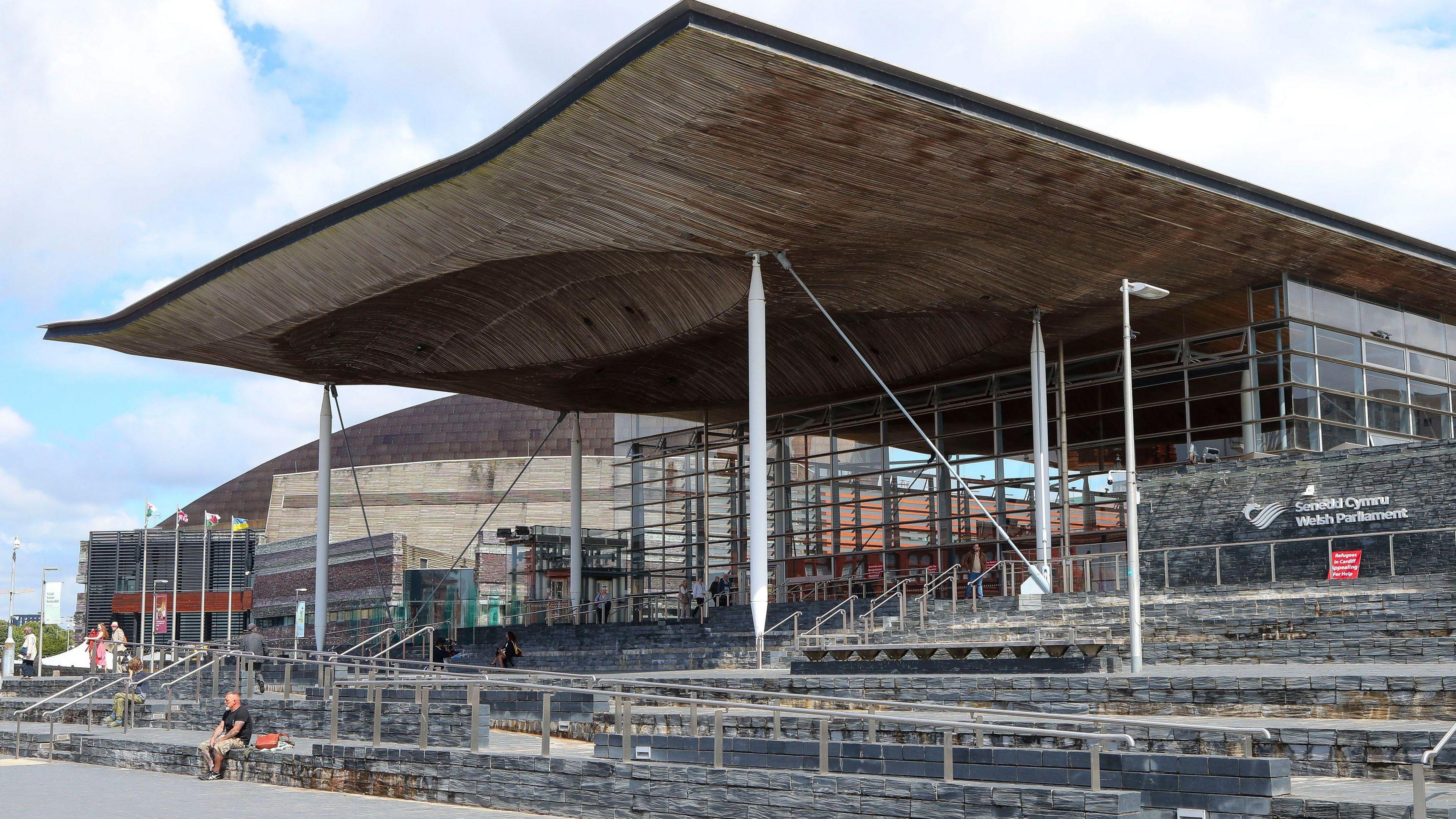 An exterior shot of the Senedd building. One man sits on the stone steps leading up to the building, and a few people are seen in the distance by the entrance.