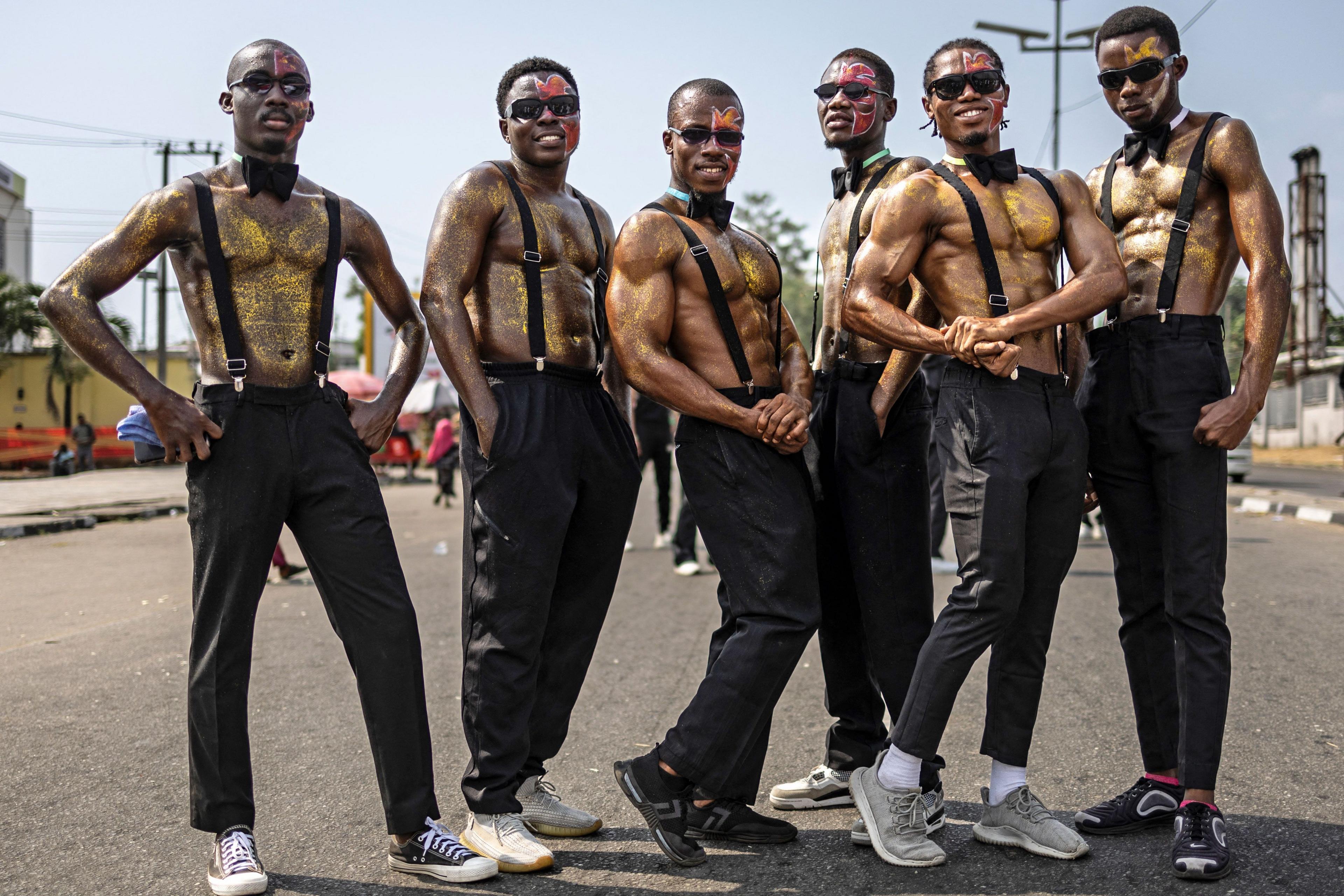 A group of men taking part in the Calabar Carnival pose for a photo in black trousers, black braces and sunglasses. Their chests are bare and covered in gold glitter. Some are wearing black bow ties.