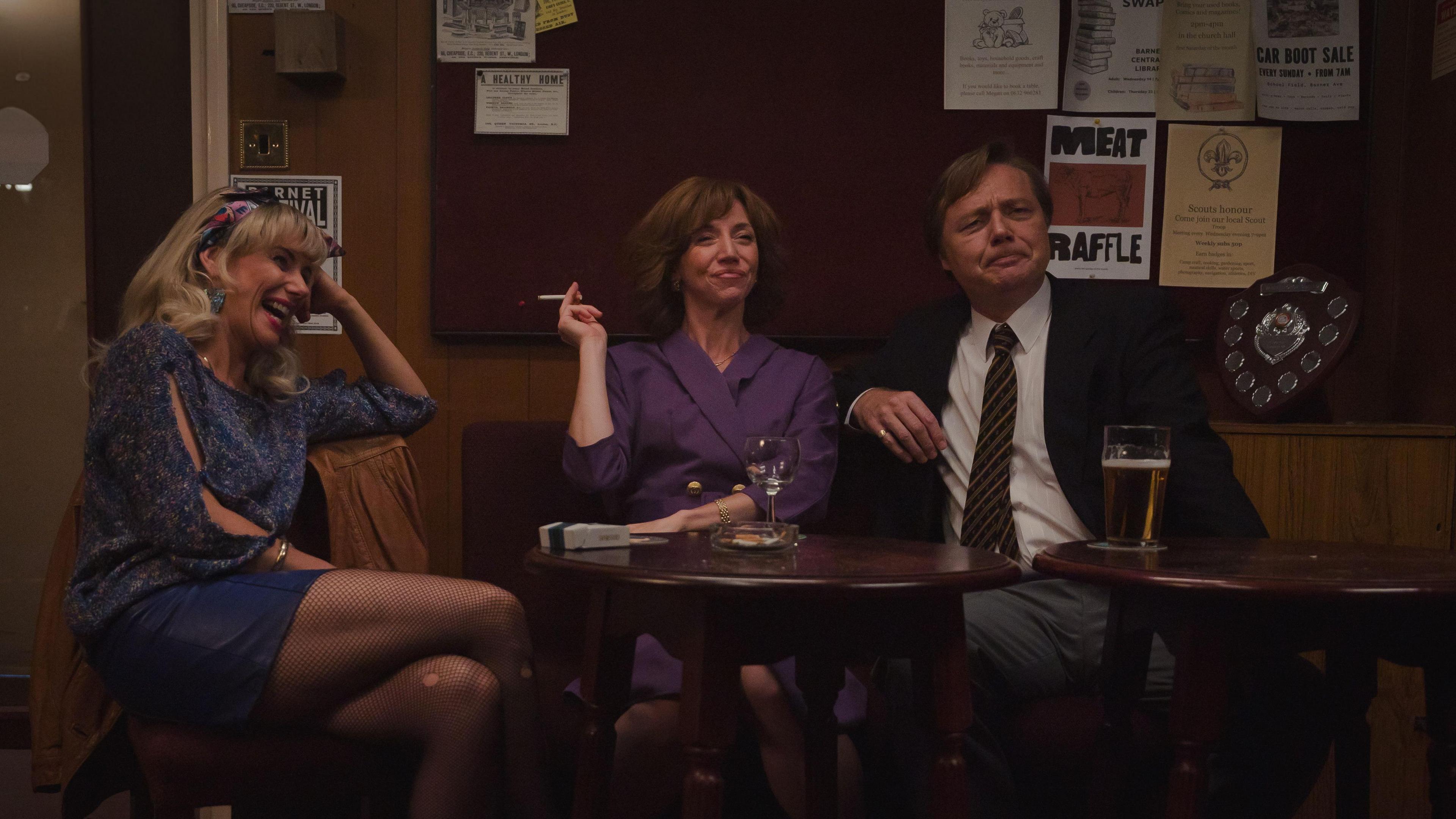 In a scene from Changing Ends, Nancy Sullivan is set behind a table in pub with a glass of wine. She has a cigarette in one hand and is wearing a purple jacket. In front of her is an empty wine glass. Shaun Dooley is to her side. Wearing a shirt, tie and black jacket. A partially drunk pint of beer is in front of him on the table. To the other side is another actress in a blue top and blue skirt. She is laughing while resting her head on her hand.