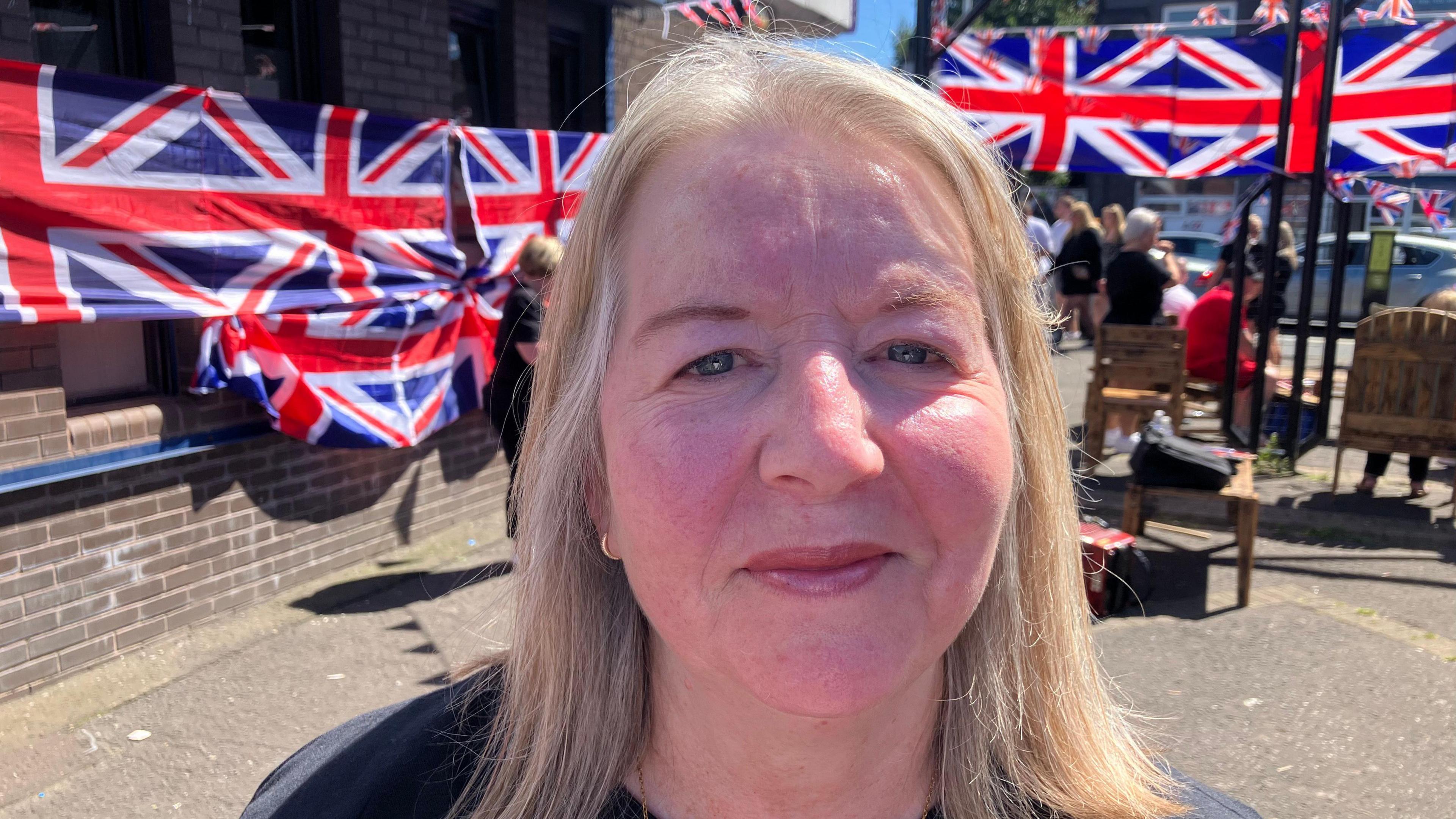 Julie Davidson has shoulder-length blond hair and wears a black t-shirt. She is standing outside and there is union jack bunting and flags in the background. 