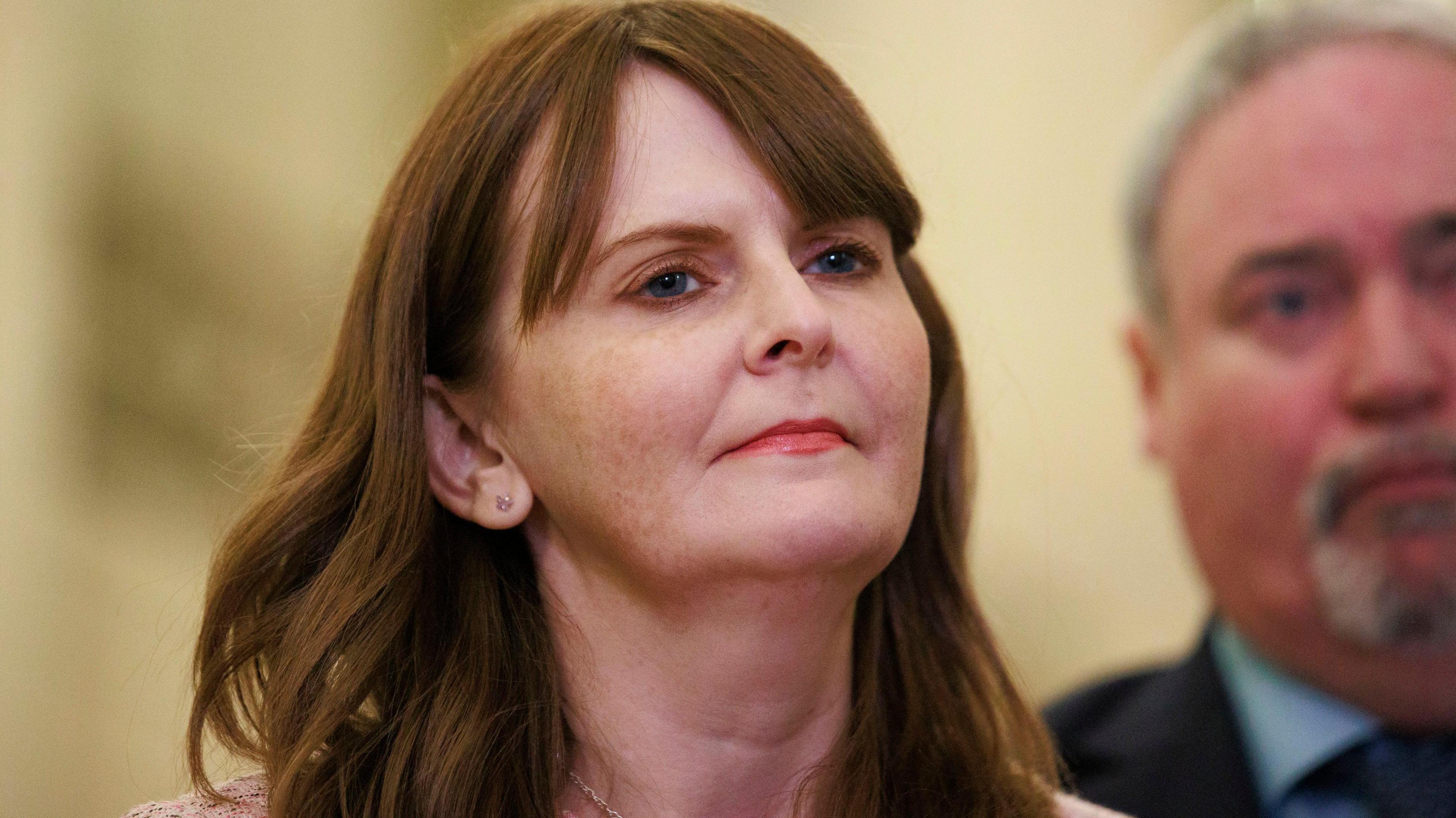 A red haired women wears red lipstick and gold earrings. She has on a pink woollen blazer. behind her left shoulder, out of focus, is a grey haired man with a grey goatee. He wears a navy suit and tie, with a blue shirt. 