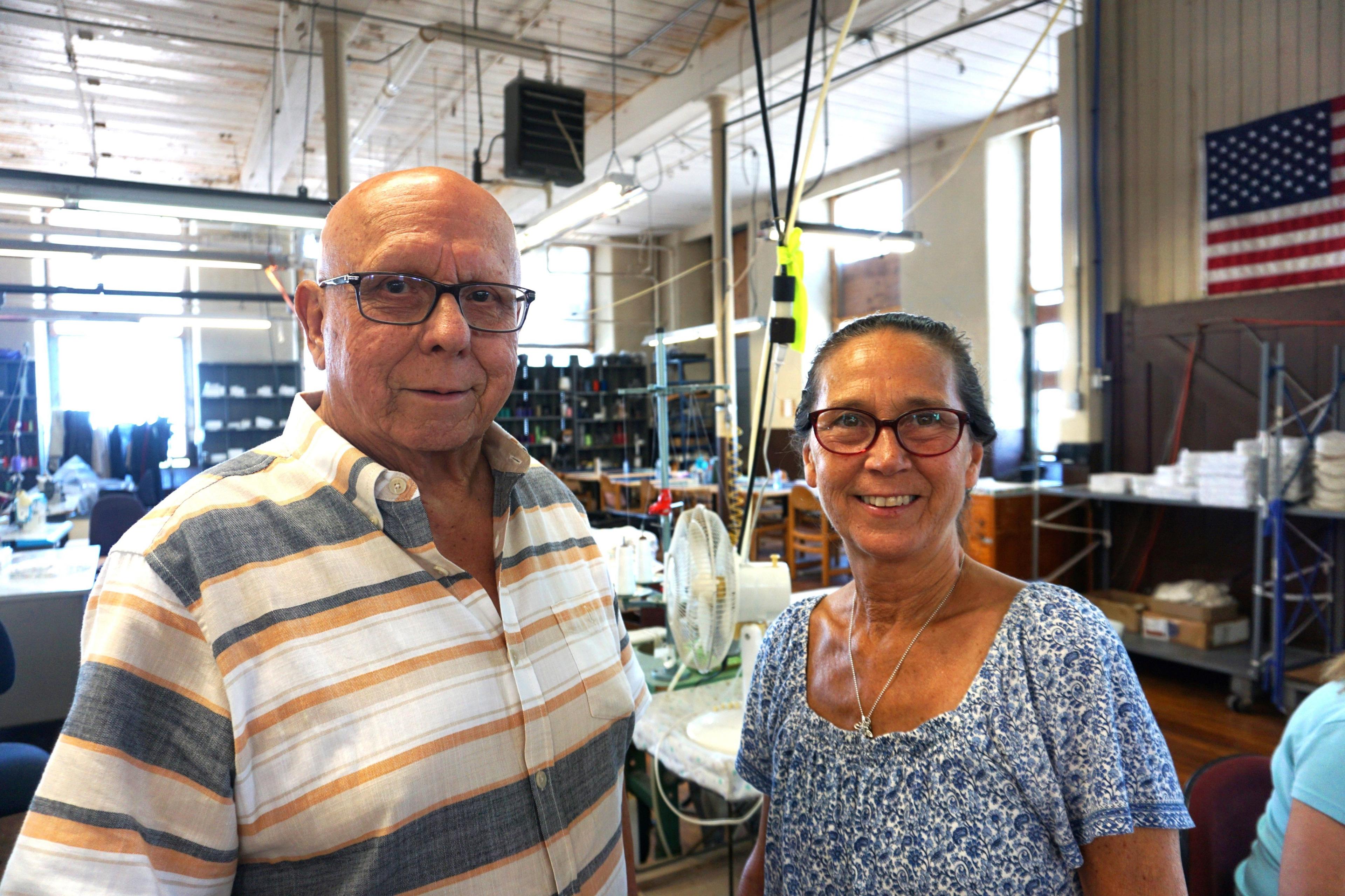 Frank and Sue stand next to each other, smiling, in front of their factory floor. Behind them is a table with a fan, and an American flag hangs from the wall.
