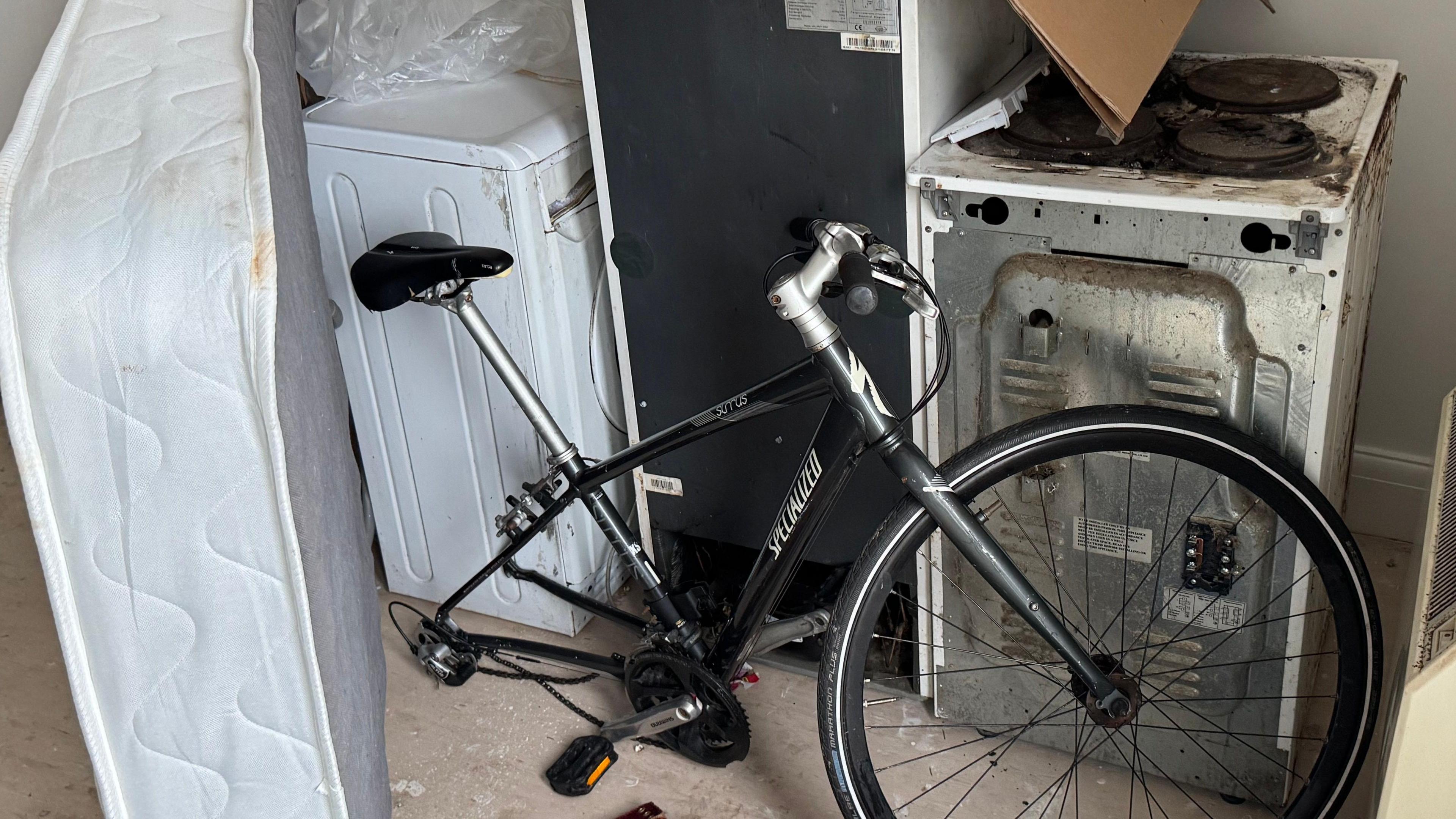 A number of white goods in Barry's bedroom. There is part of a black bike, a defunct oven, washing machine, fridge and mattress.