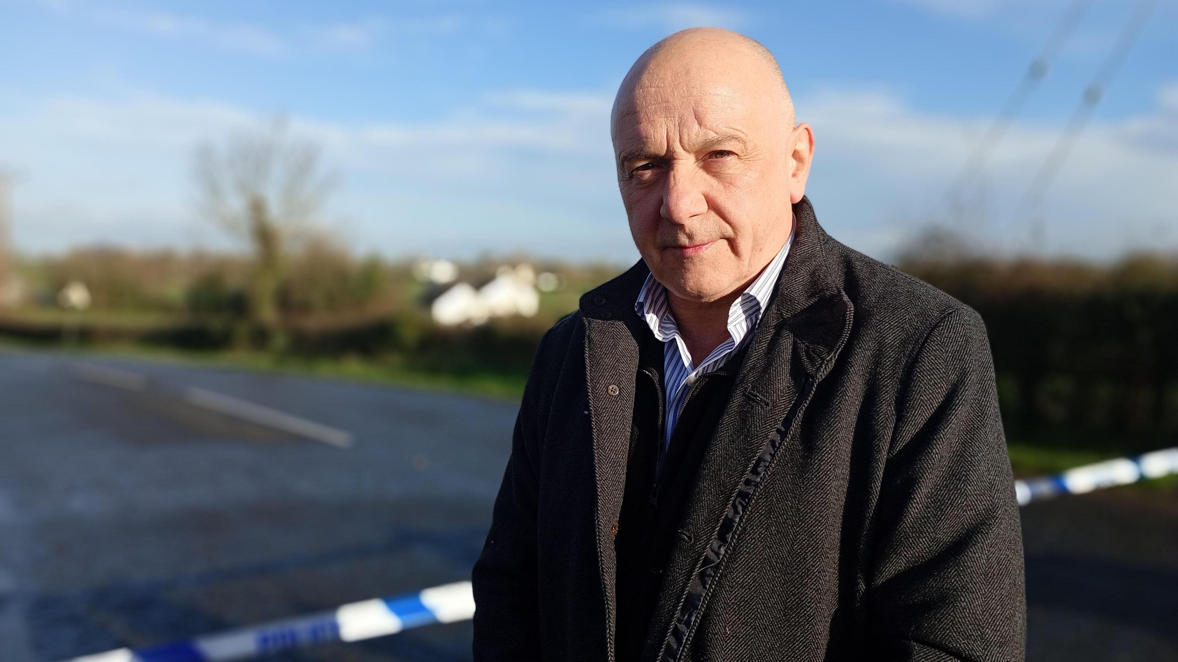 A bald man wearing a black coat over a blue and white striped shirt is standing in front of a rural road which is closed off by a blue and white police cordon. The background is out of focus but a hedgerow, tress and white building can be seen.