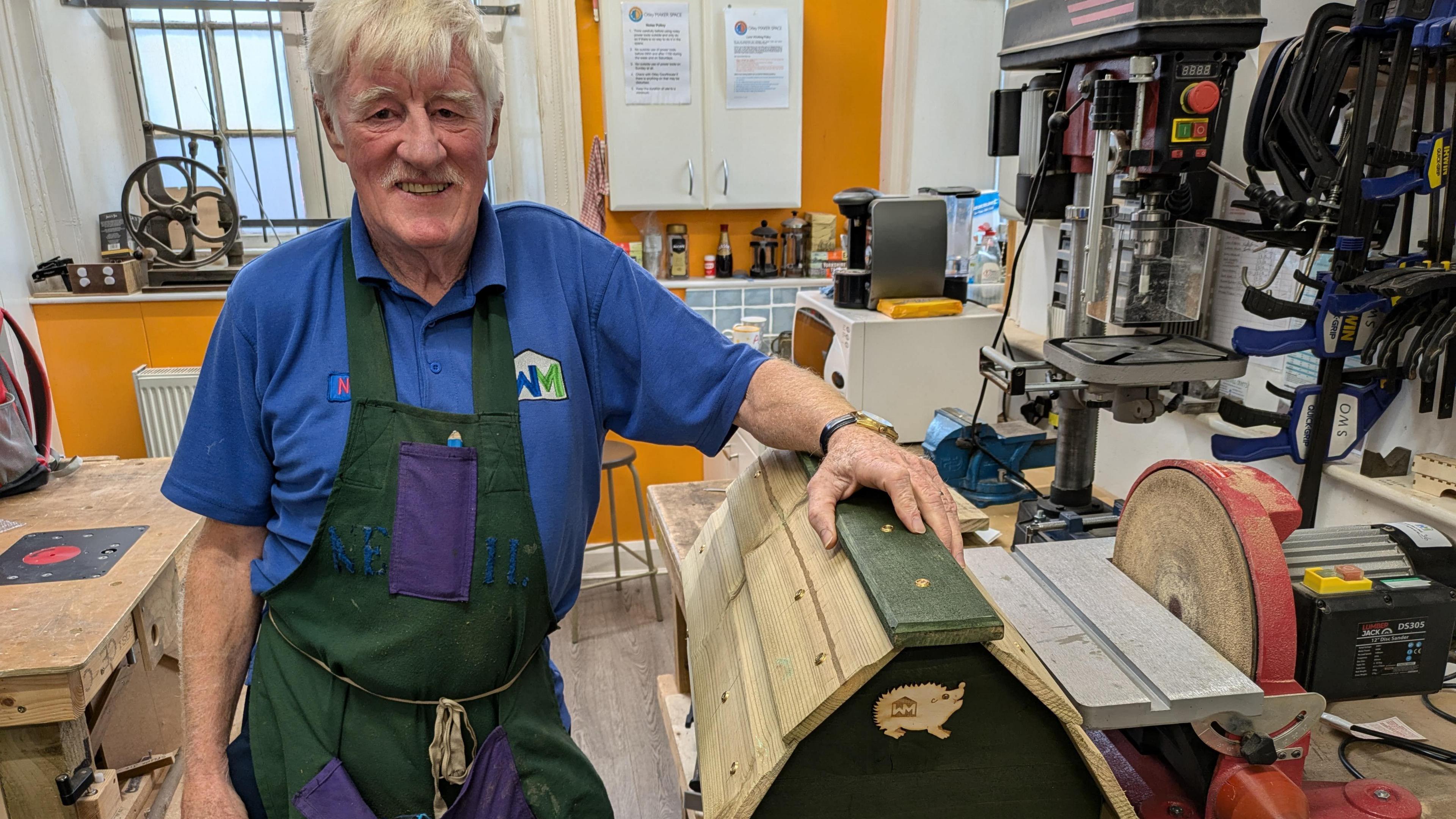 A gentleman in his seventies smiles into the camera with his left hand perched on a wooden crate, which has a small wooden cut-out of a hedgehog at the front. He is standing in a workshop with tools and machinery all around him.