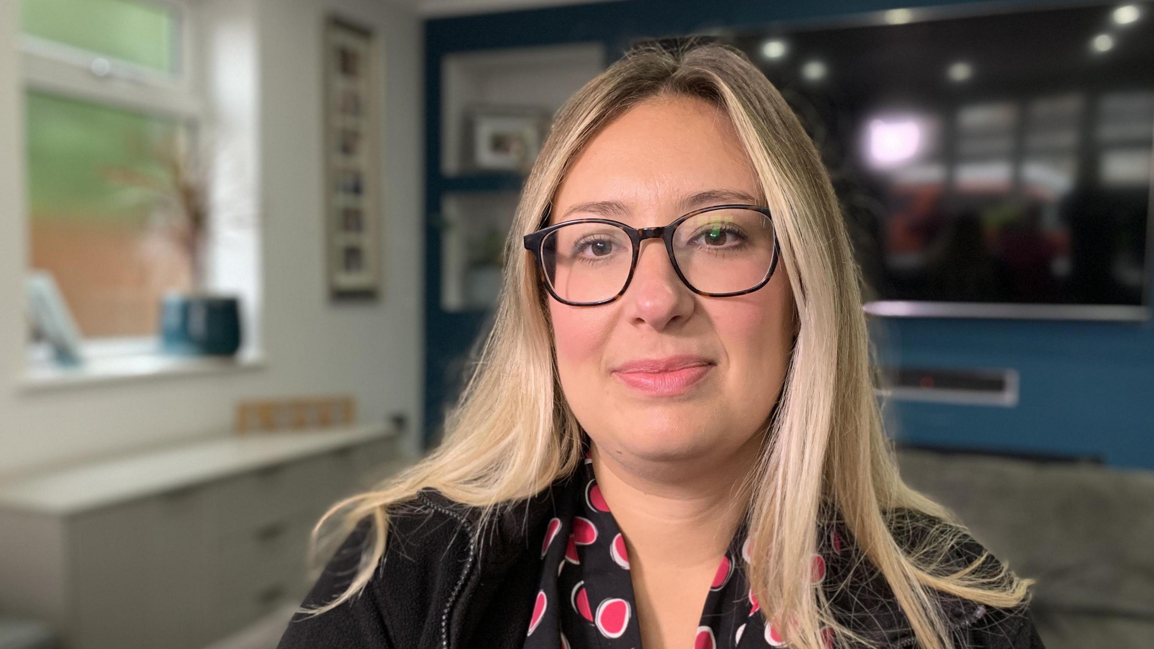 A 34-year-old woman is wearing a black cardigan and a blouse with pink spots. She is also wearing glasses and has long, blonde hair.