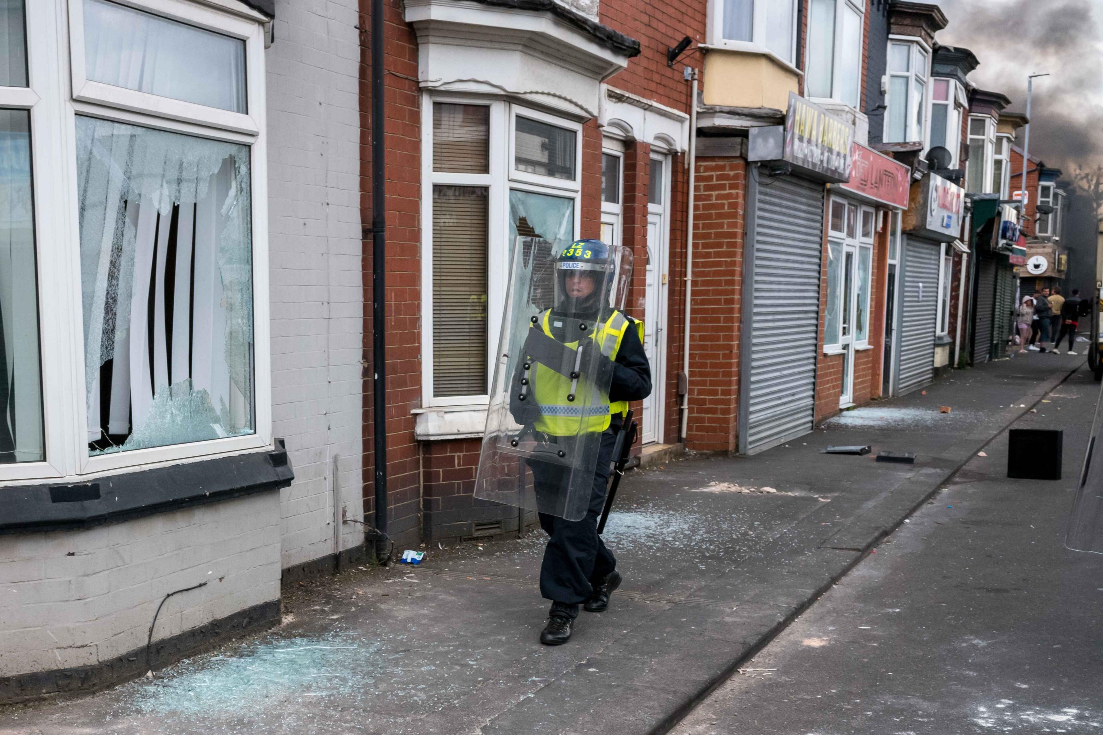 A police man holding a riot shield stands in front of a house with smashed windows during a riot
