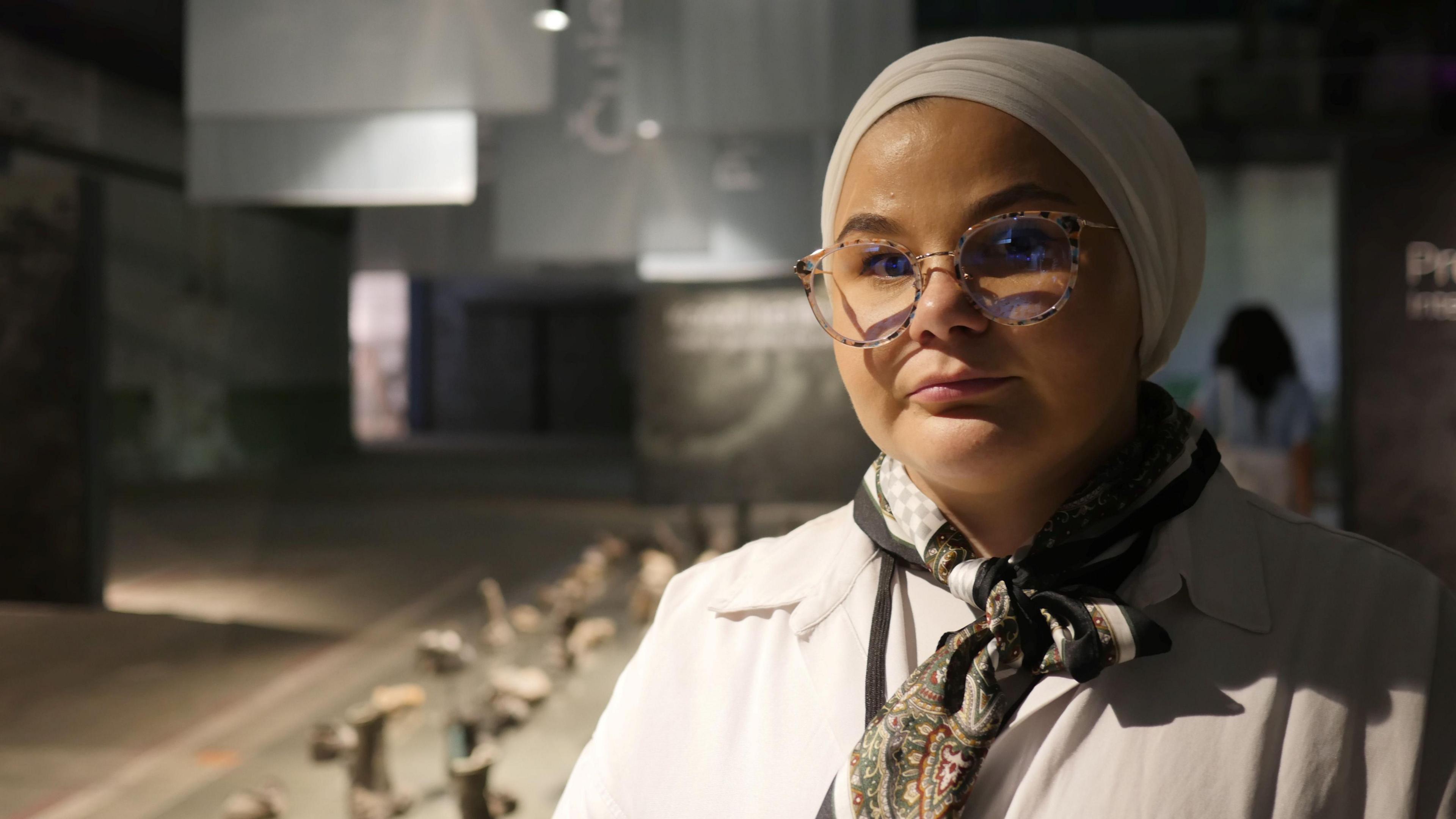 A woman wearing glasses, with her hair covered by a scarf looks directly into the camera. She is wearing a white coat and a scarf. Behind her is the Srebrenica Memorial Centre, a grey exhibition space displaying the shoes of victims of the massacre.