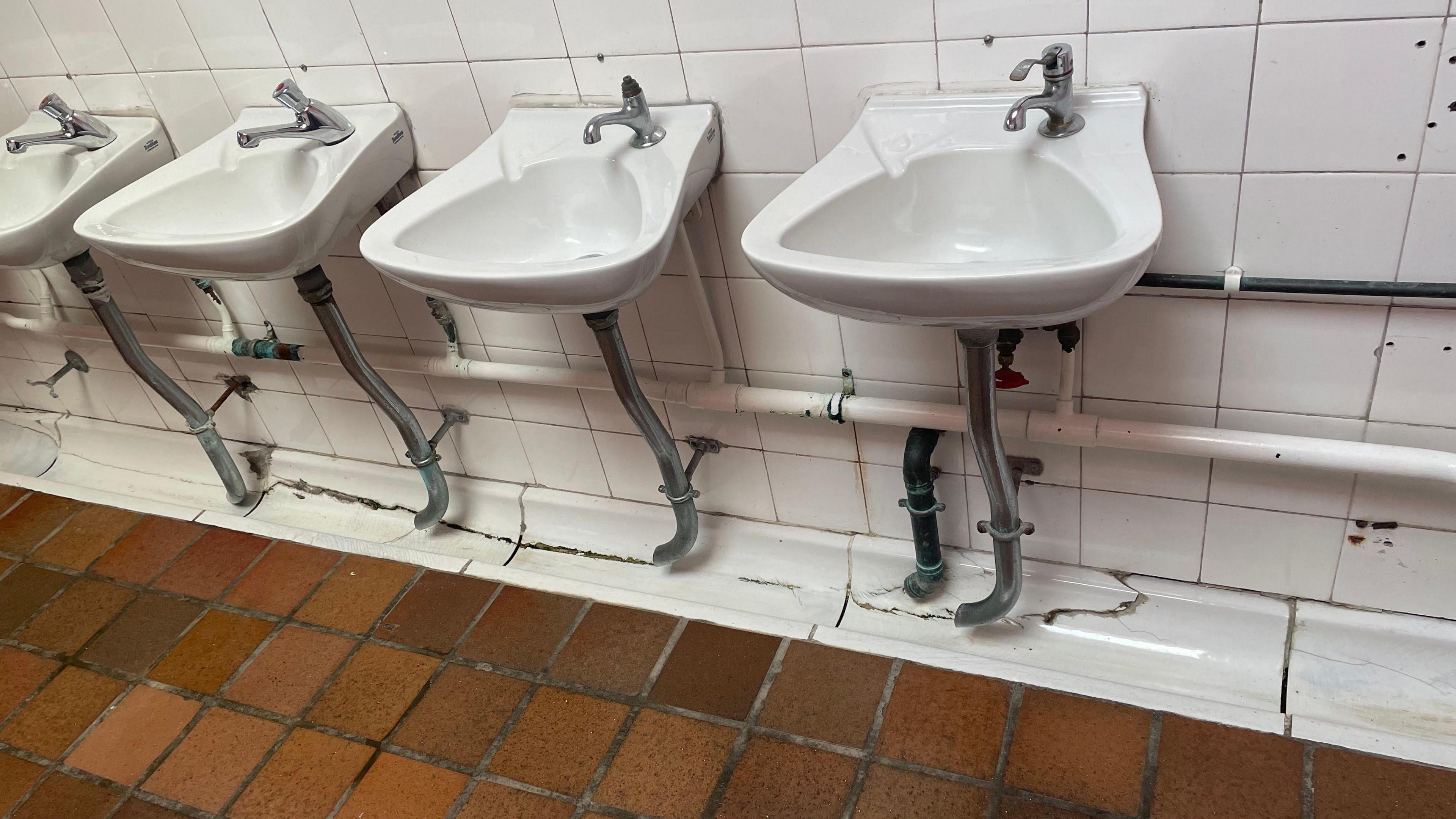 A tiled bathroom. The tiles on the ground are coloured like bricks while the wall tiles are white. The sinks are white. The tiles are stained and cracked in places.