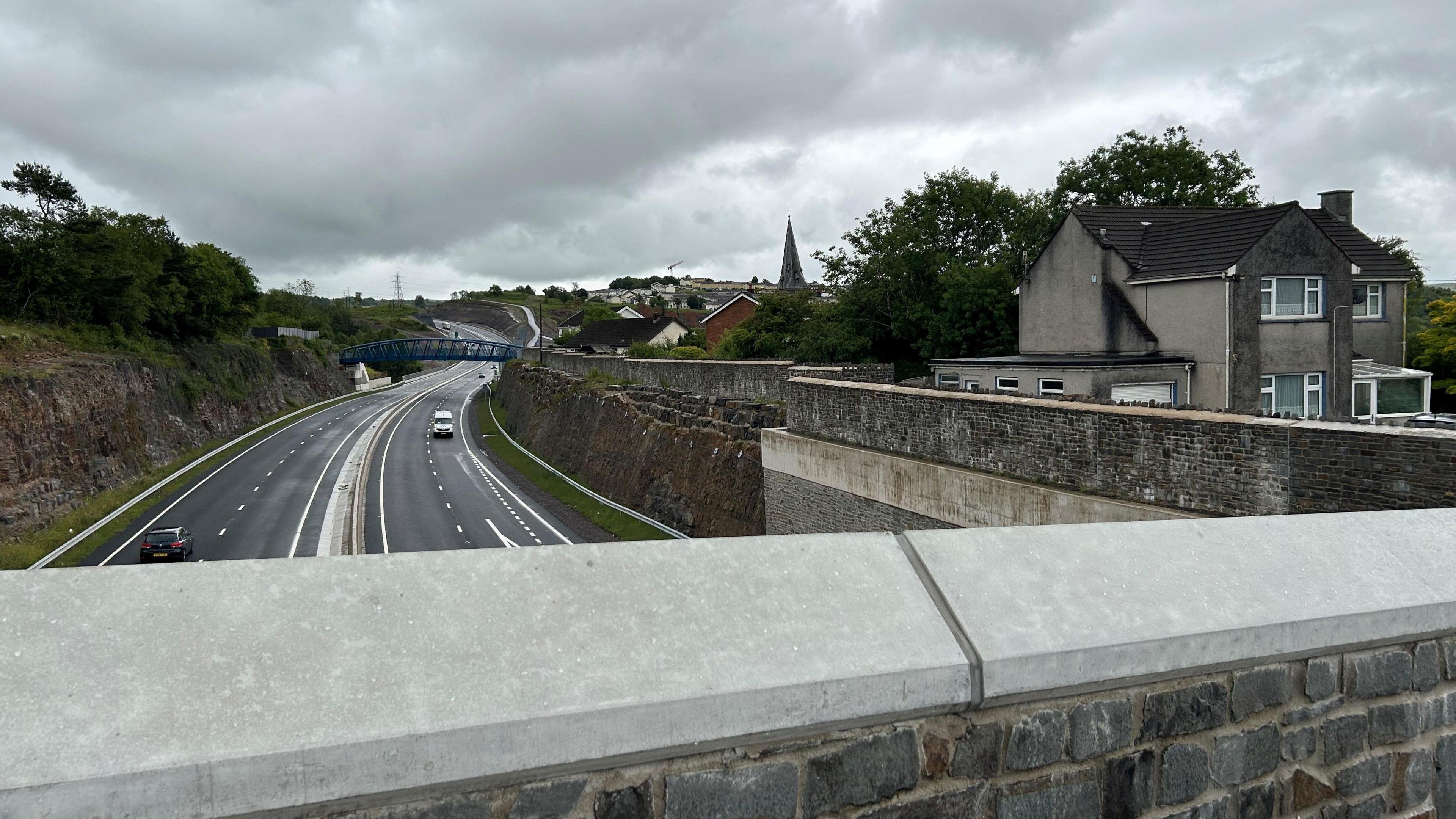 A view from a bridge down onto the Heads of the Valleys dual carriageway. On the right hand side is a large stone wall, and John's house on a platform to the right of the wall. It is a cloudy day.