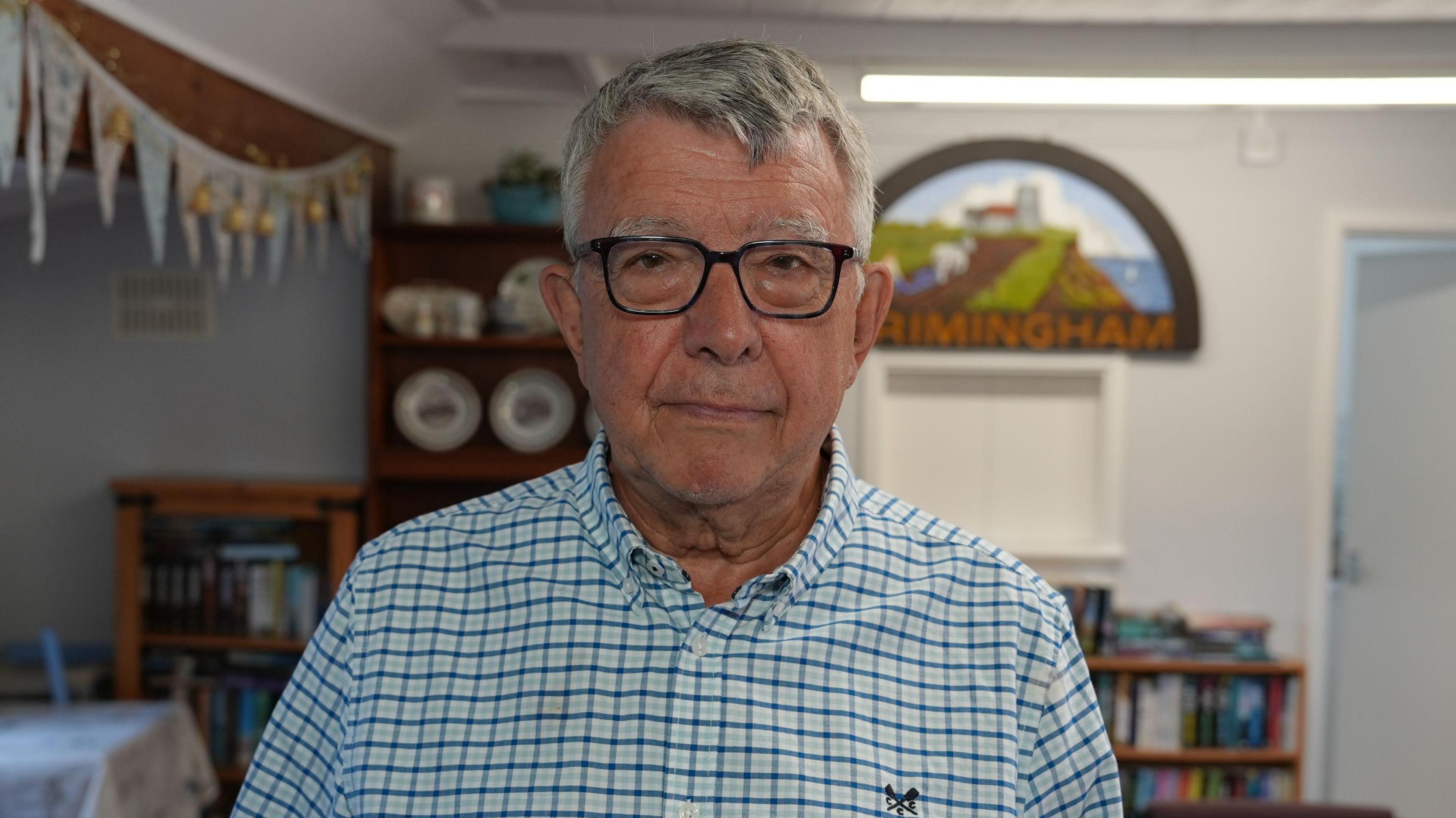 Terry Brown, a man standing inside Trimingham village hall. He is looking directly at the camera and is wearing a blue and white checked shirt and a pair of black frame glasses on his face. In the background is a sign hanging on a white wall which says TRIMINGHAM on it. To the left is a row of bunting hanging up across a wooden beam and there is a wooden shelving unit behind him with a range of crockery on it.