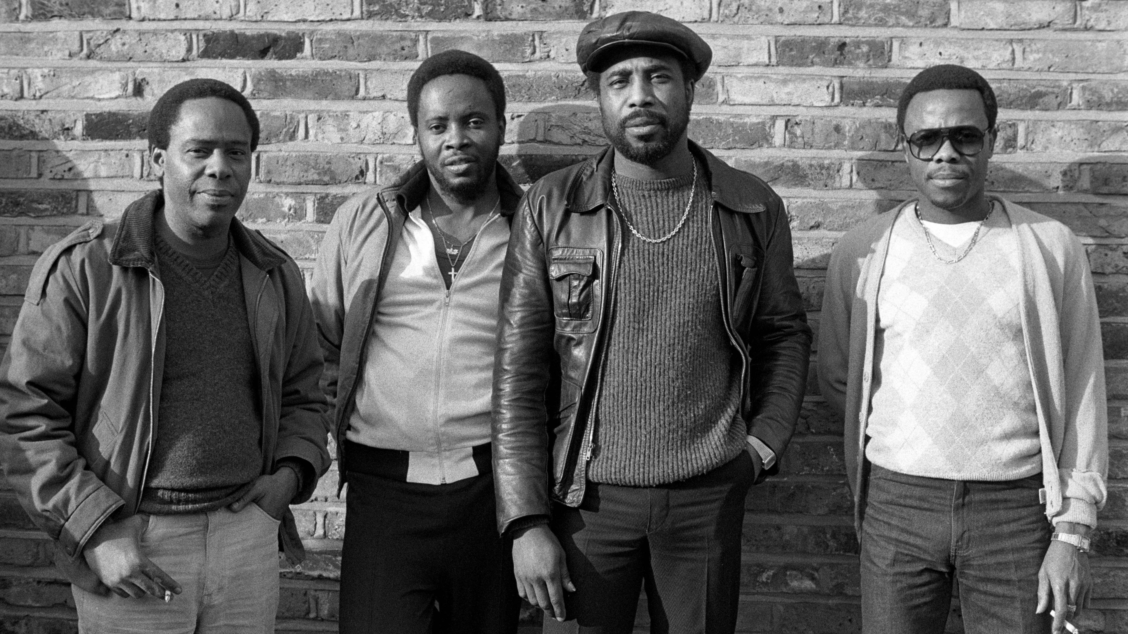 Black and white photo of B.B. Seaton of The Gaylads, Jackie Robinson of The Pioneers, A J Franklin of Chosen Few and Junior English standing in front of a brick wall