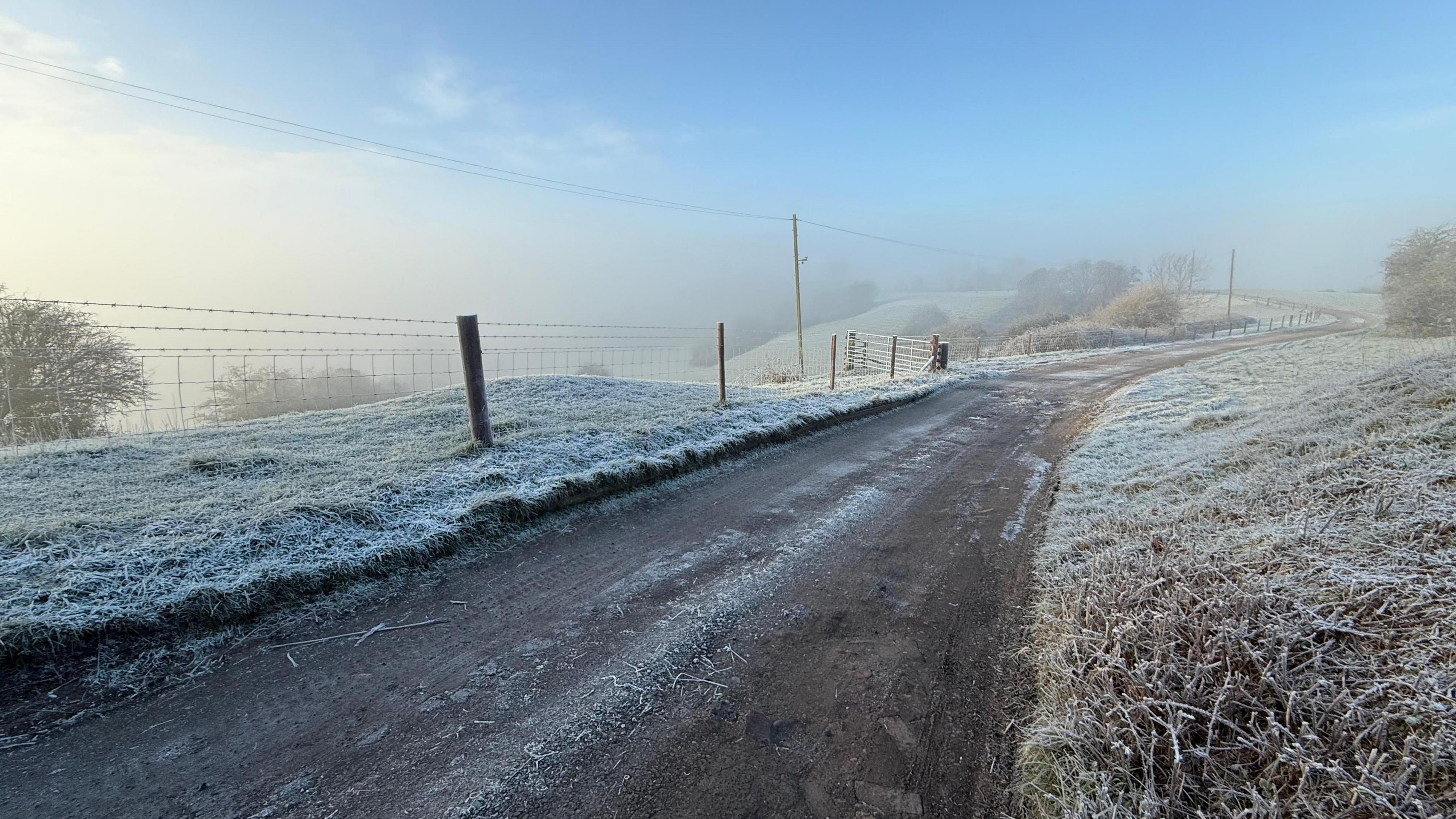 A road is running through frosty green fields with a blue sky overhead. Fog has gathered to the left of the sky, while the right side has minimal clouds.