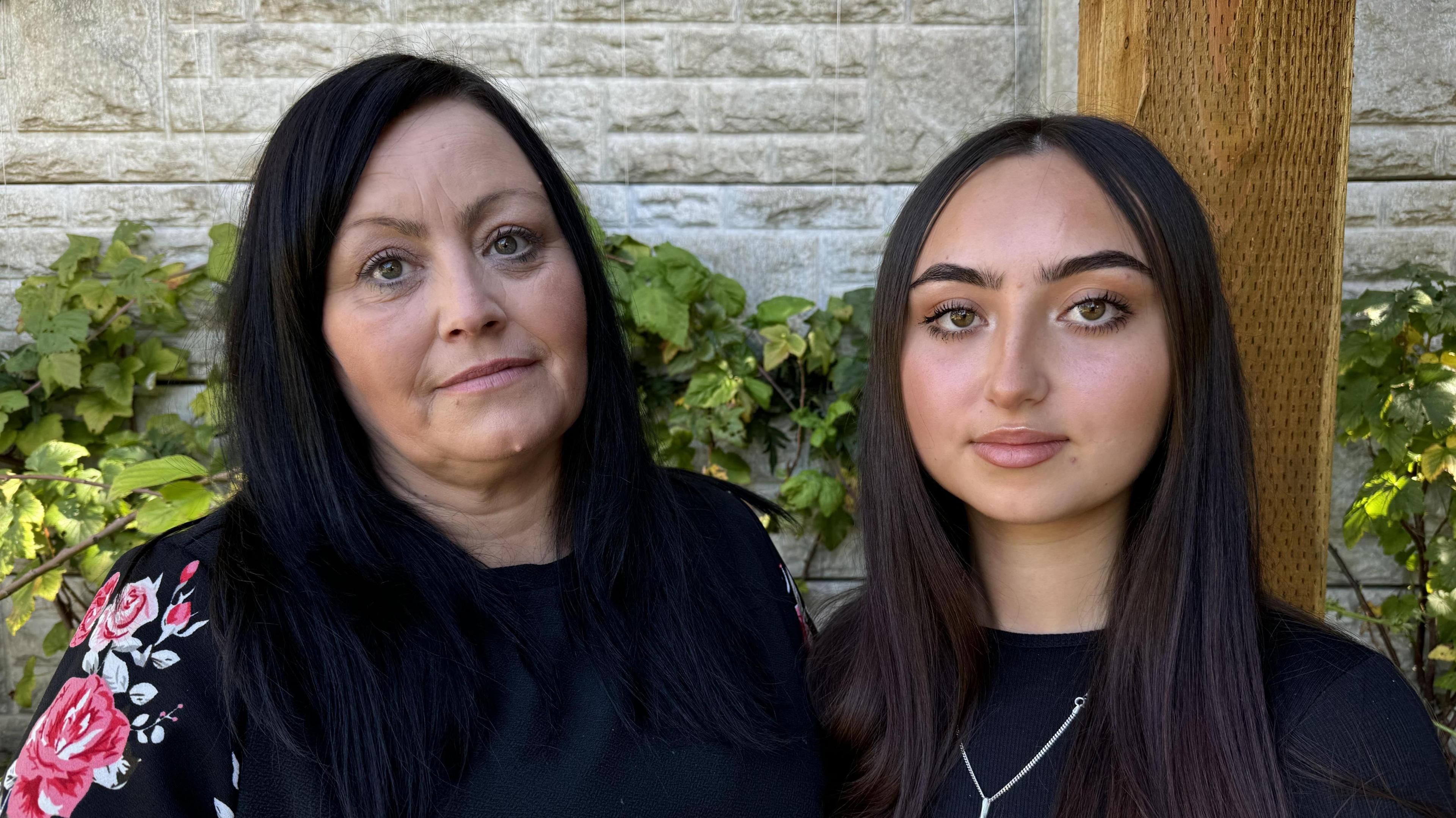 Celyn and her mum Emma stood next to each other. There is a grey wall behind them with some green leaves creeping up it.