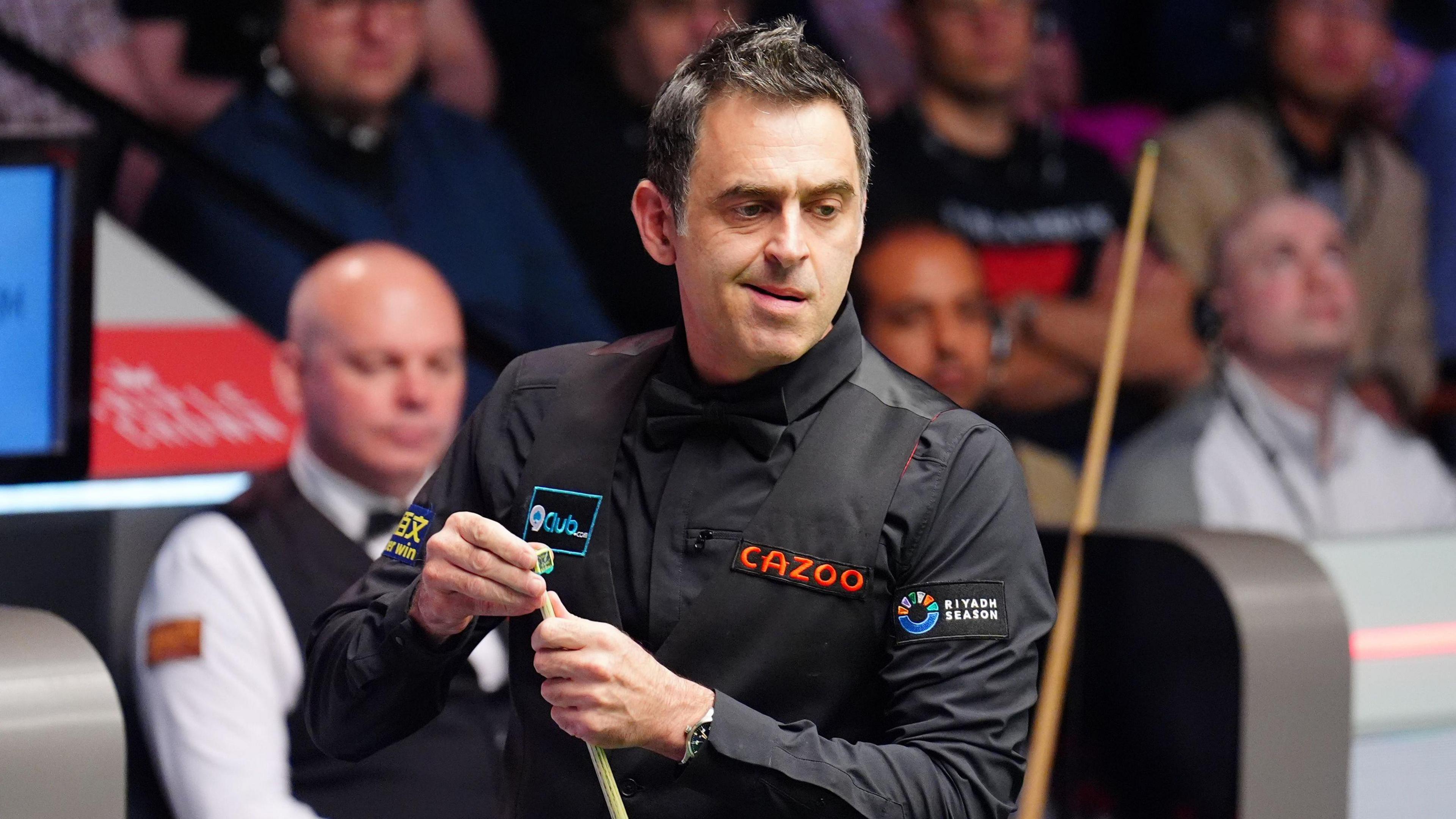 Ronnie O'Sullivan applying chalk to his snooker cue. His competitor and the crowd behind him are out of focus.