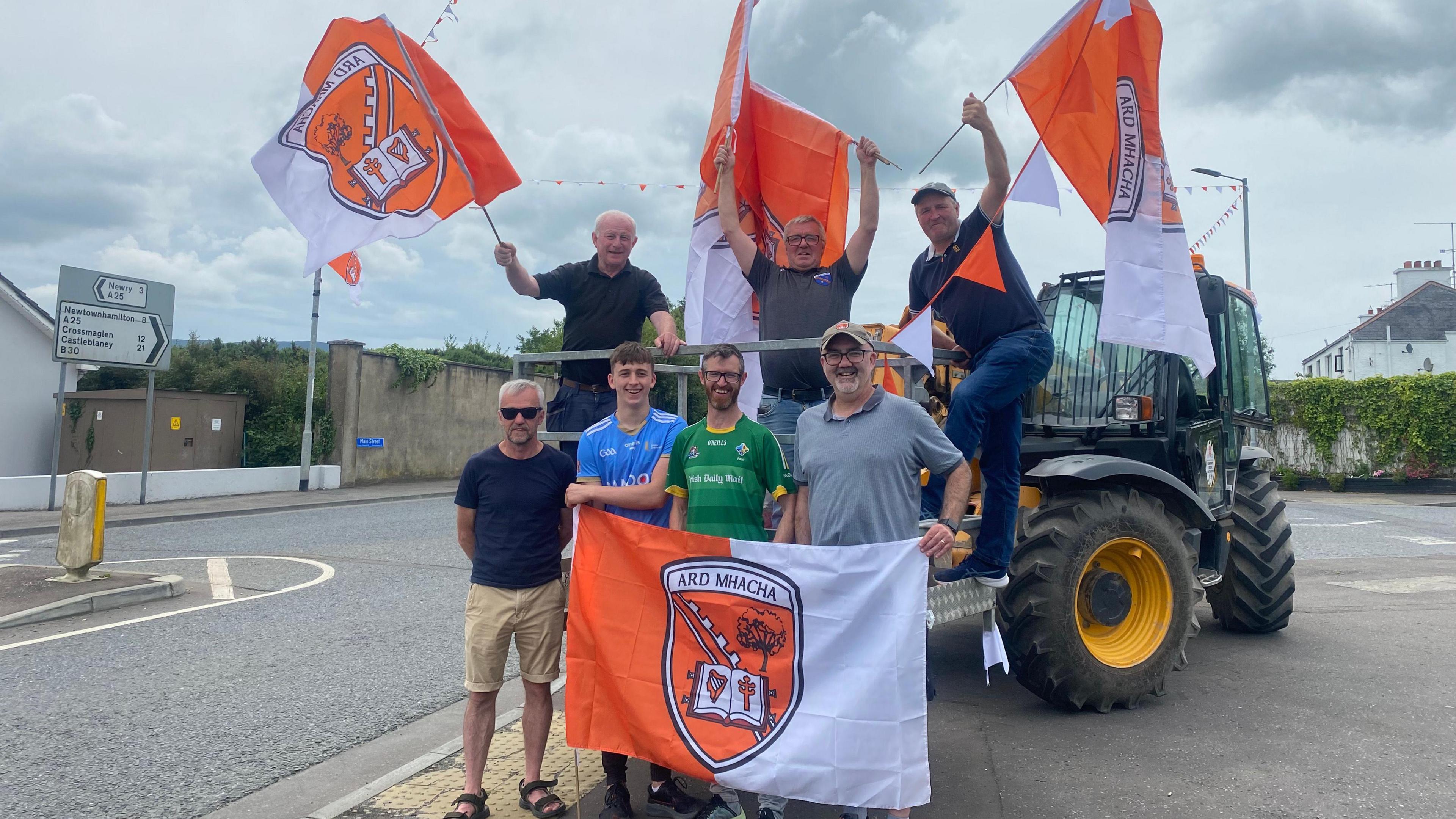 A group of seven men are standing on a trailer attached to a green tractor. They're holding orange and while flags with the Armagh GAA crest