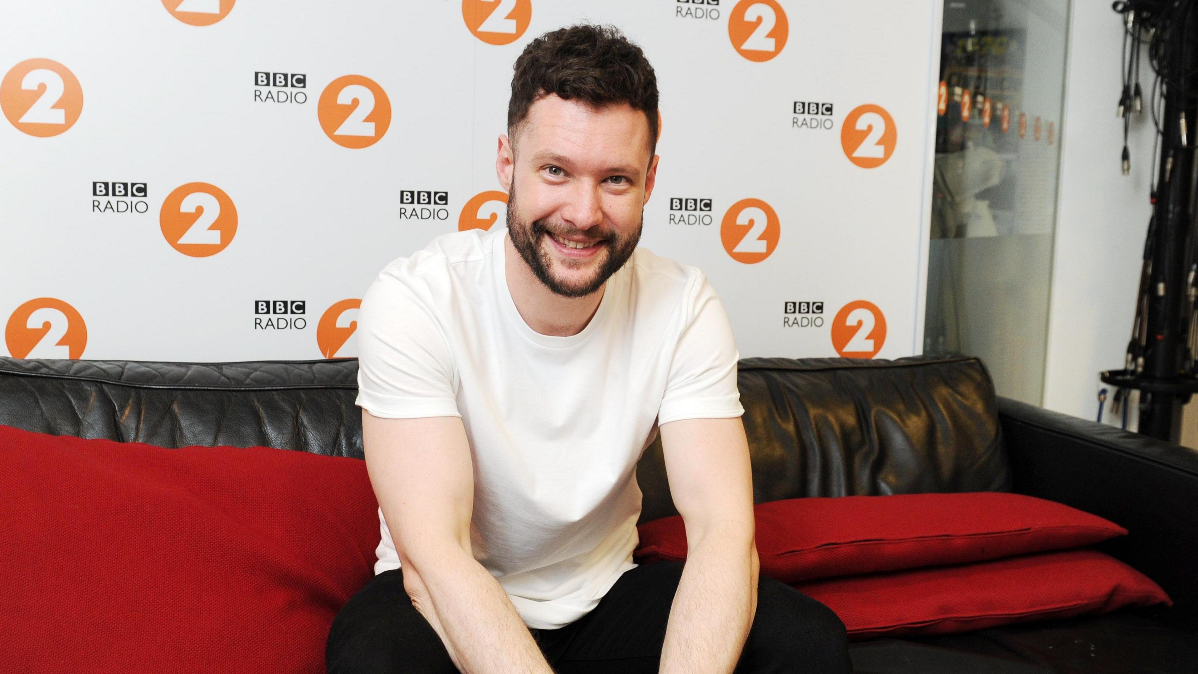 Calum Scott, wearing a white t-shirt and black jeans, sat on a sofa at Radio 2.