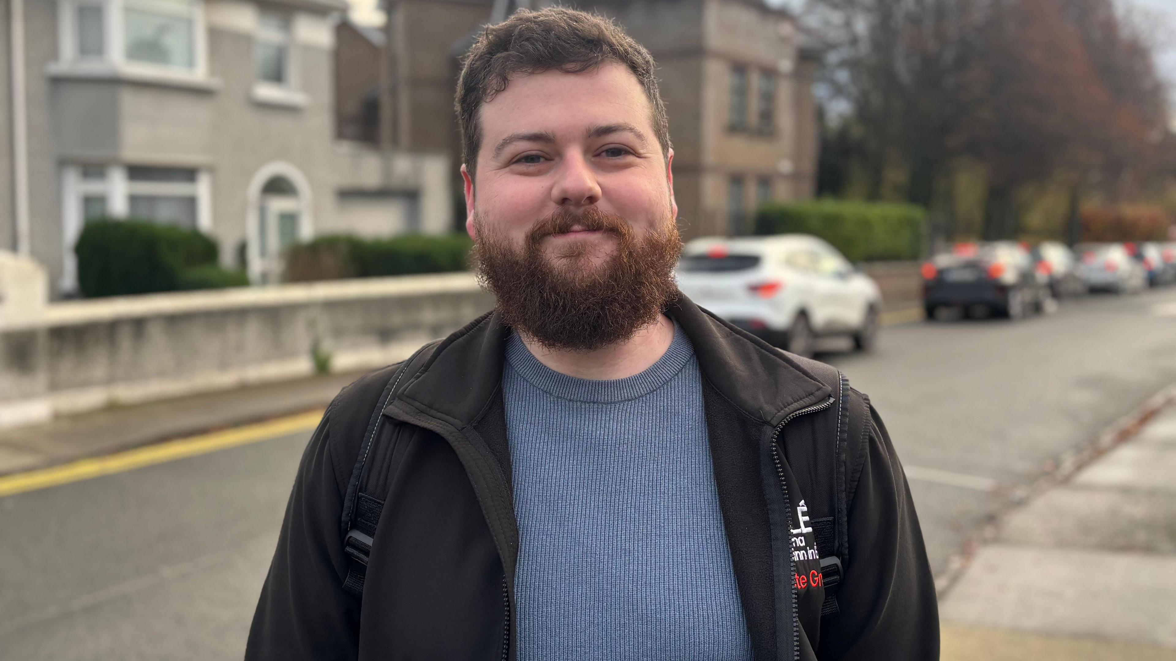 A young man who is a student union representative wearing a black jacket and a blue jumper smiles at the camera