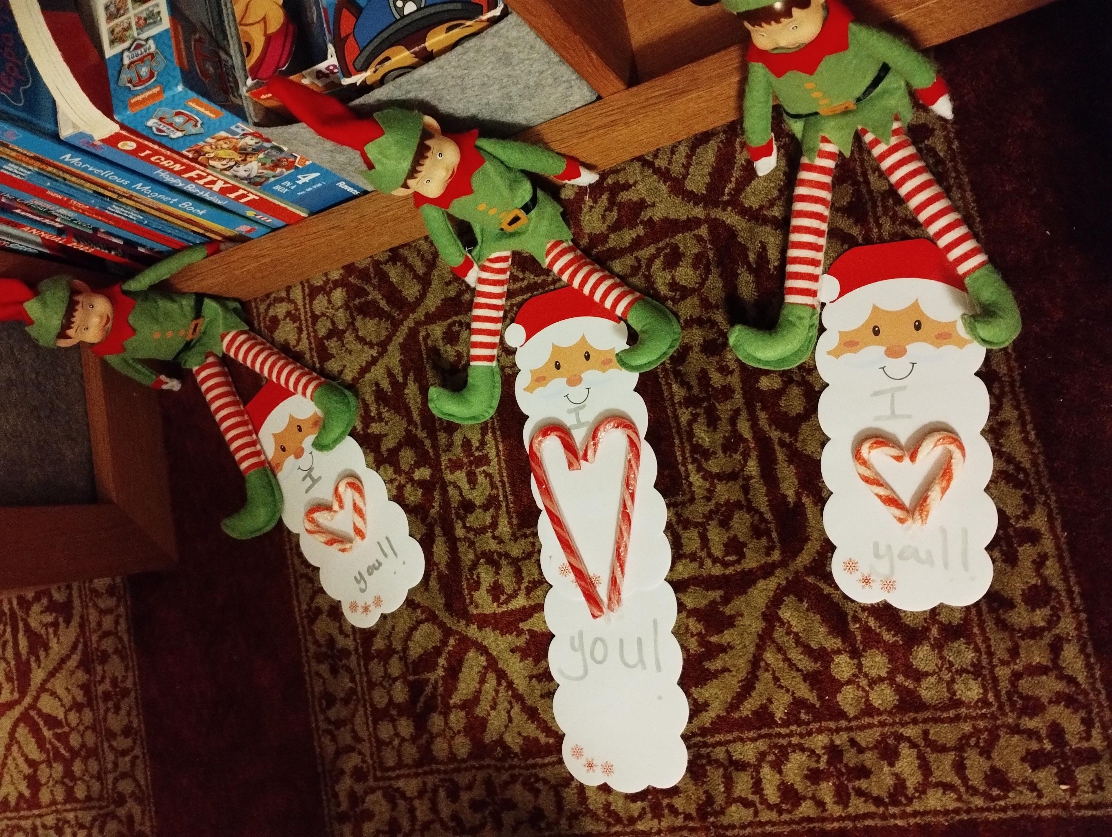 Three elves are sat on the floor. They have red and white stripey legs and wearing a green tunic, shoes and a red and green hat. Candy canes are shaped into hearts and santa cards say "I heart you".