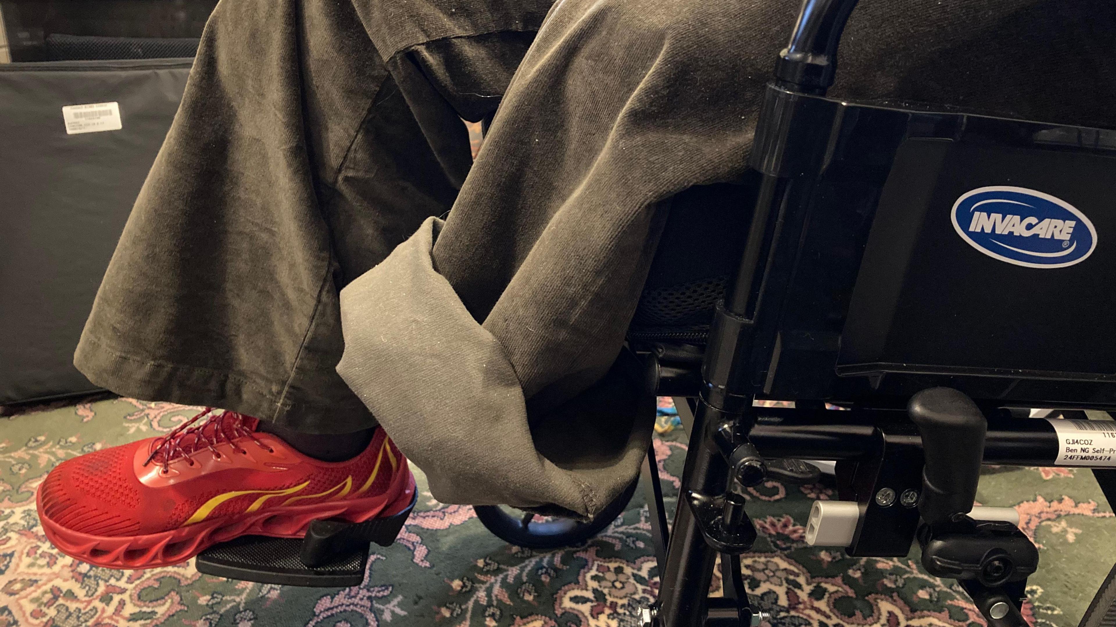 A close-up of a person sitting in a wheelchair. They are wearing grey trousers with their right foot in a red trainer. The bottom of their left trouser leg is rolled up, with no foot at the end.