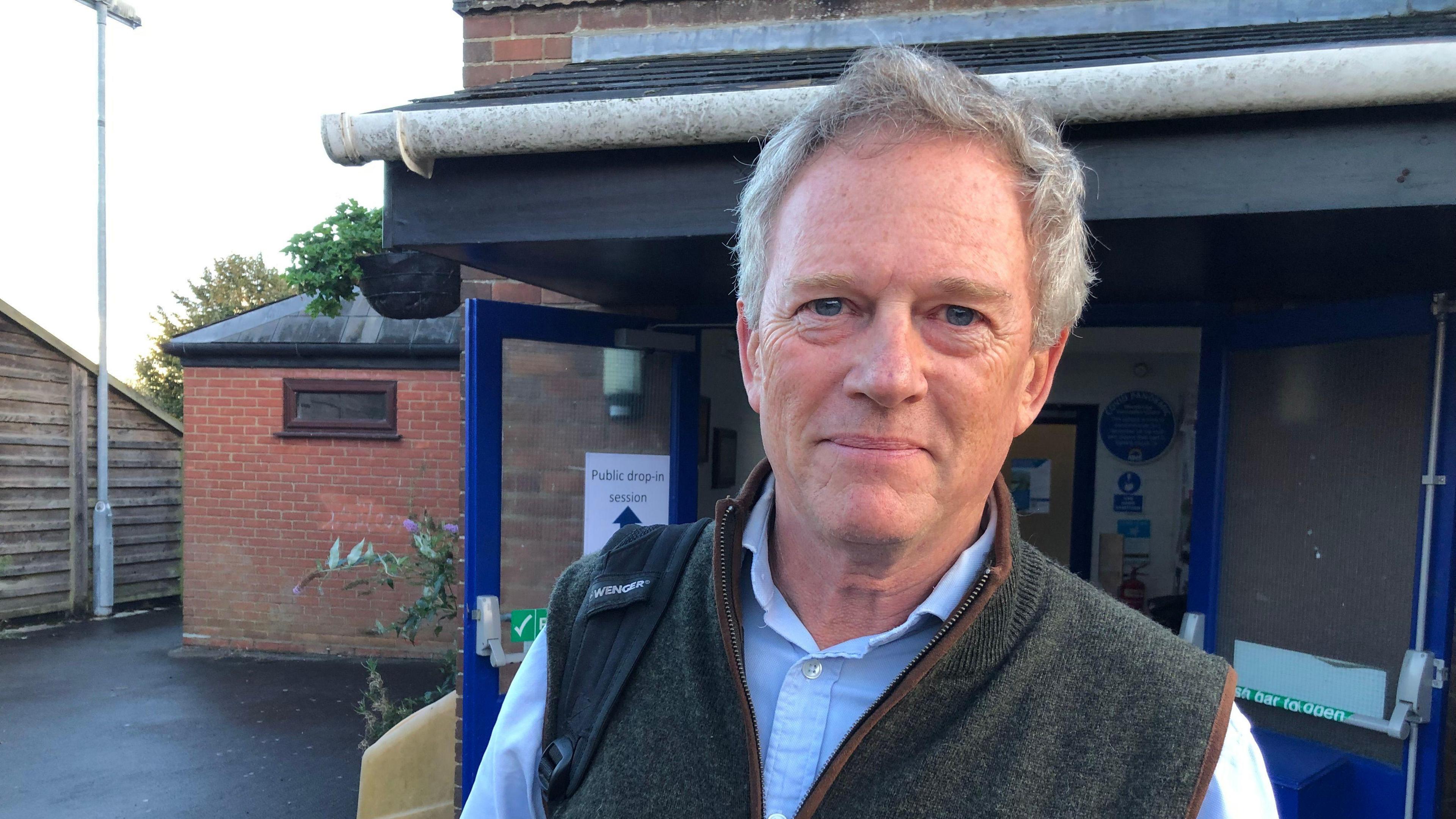 A picture of Phillip Ainsworth. He is smiling to the camera outside a community centre. He hair grey hair and is wearing a blue shirt with a tweed jacket over the top with no sleeves. A rucksack strap can be seen resting on his left shoulder.