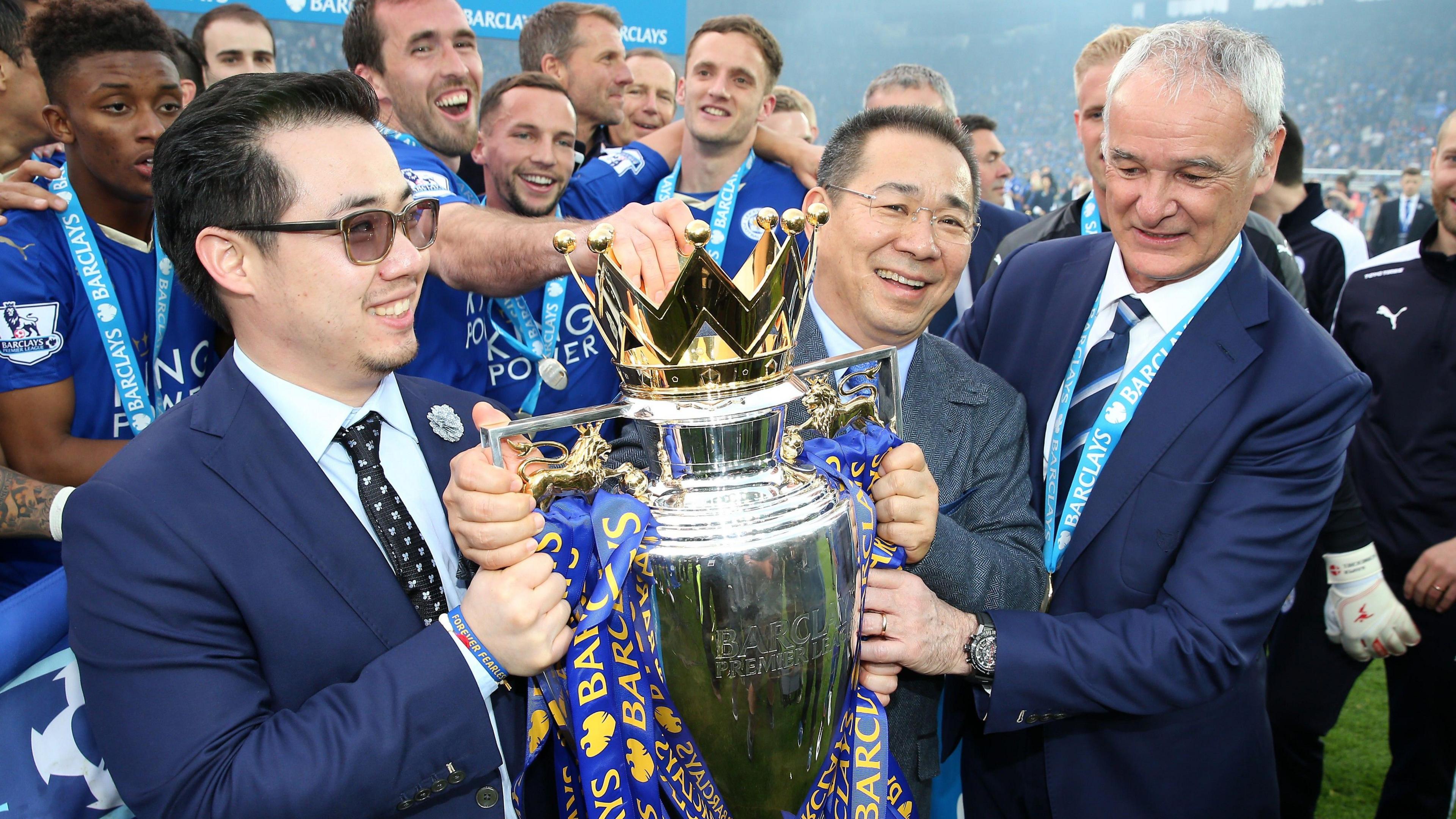 Vichai Srivaddhanaprabha stands on the King Power Stadium pitch holding the Premier League trophy with Claudio Ranieri and son Aiyawatt with the squad celebrating behind them. 