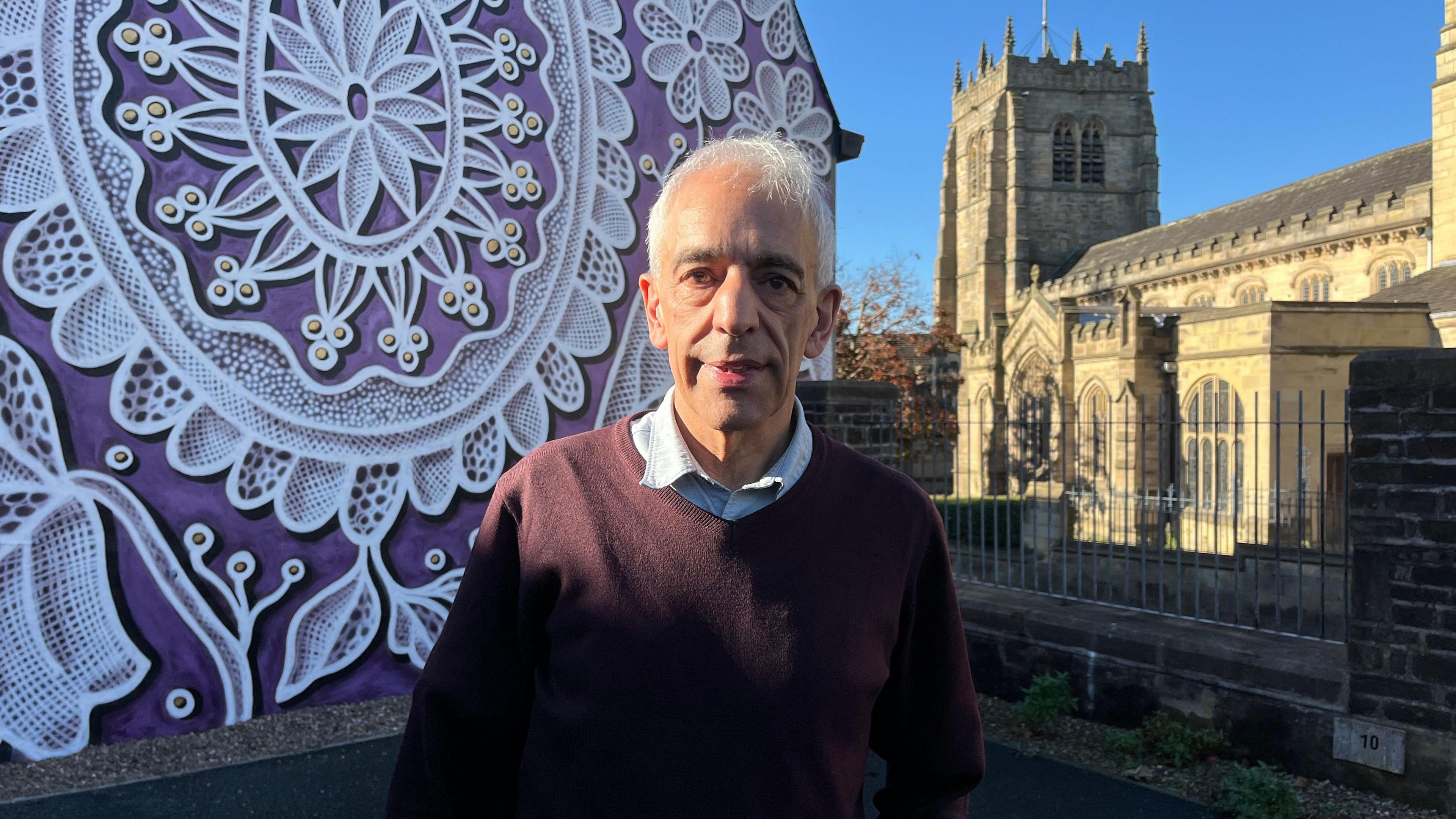 Asadour Guzelian stands in front of a white and purple floral mural with Bradford Cathedral behind him.