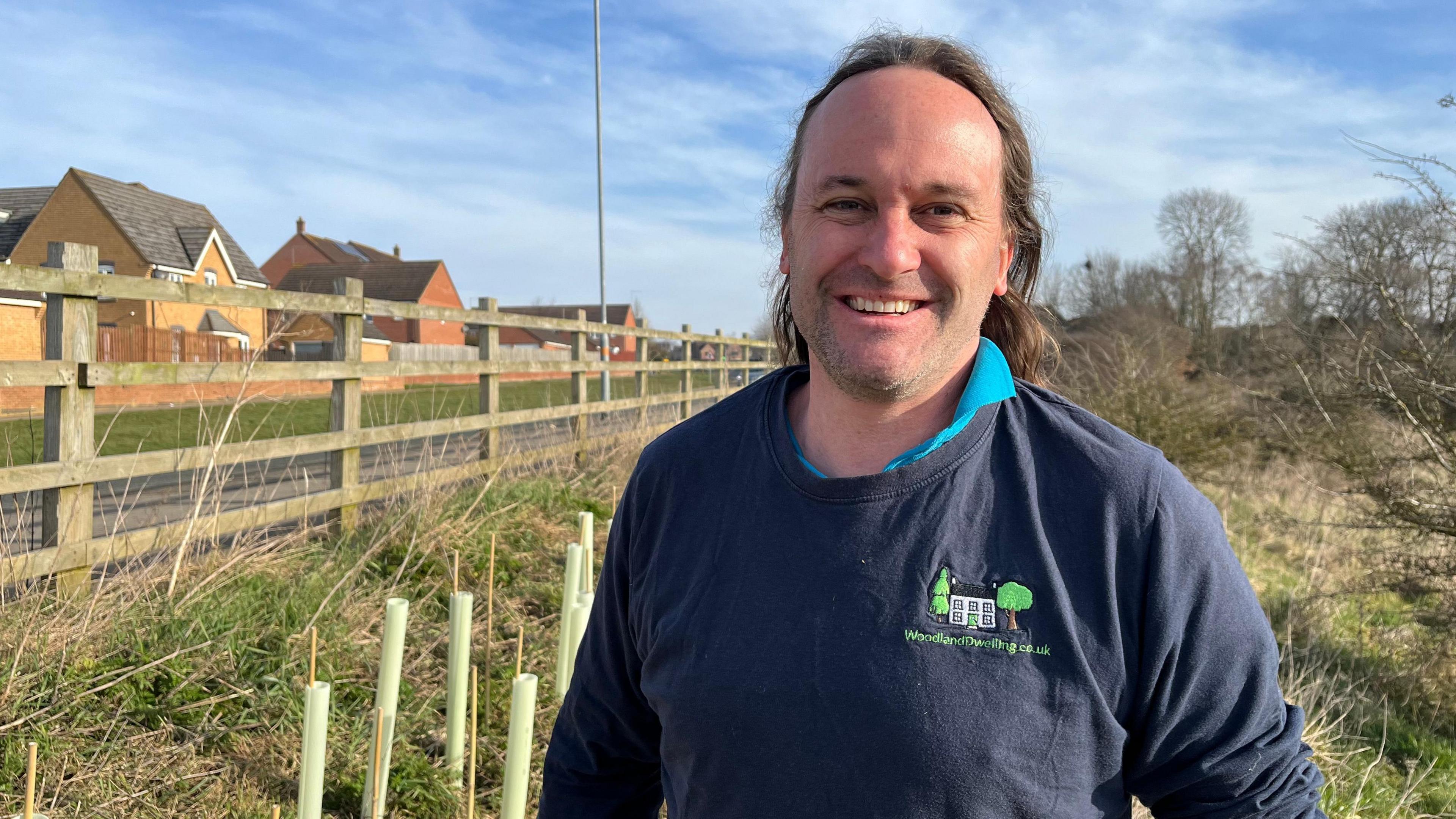 A picture of a man wearing a navy-coloured jumper with 'Woodland Dwelling' branding. He is standing behind a wooden fence with freshly-planted trees behind him. 