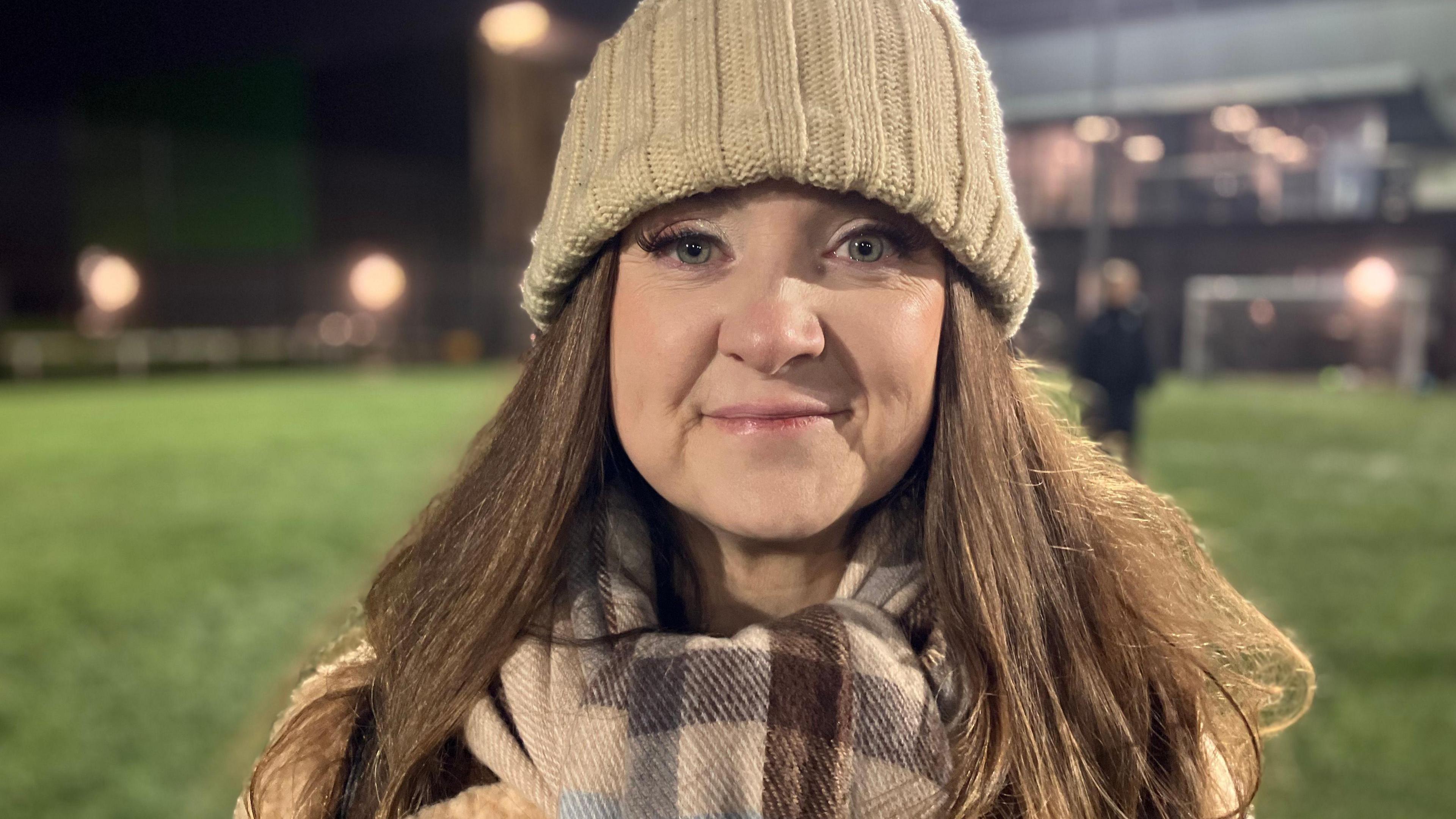 Cameron's mum Kelly Mills looking at the camera smiling. She is wearing a brown hat and scarf and there is a football pitch in the background.