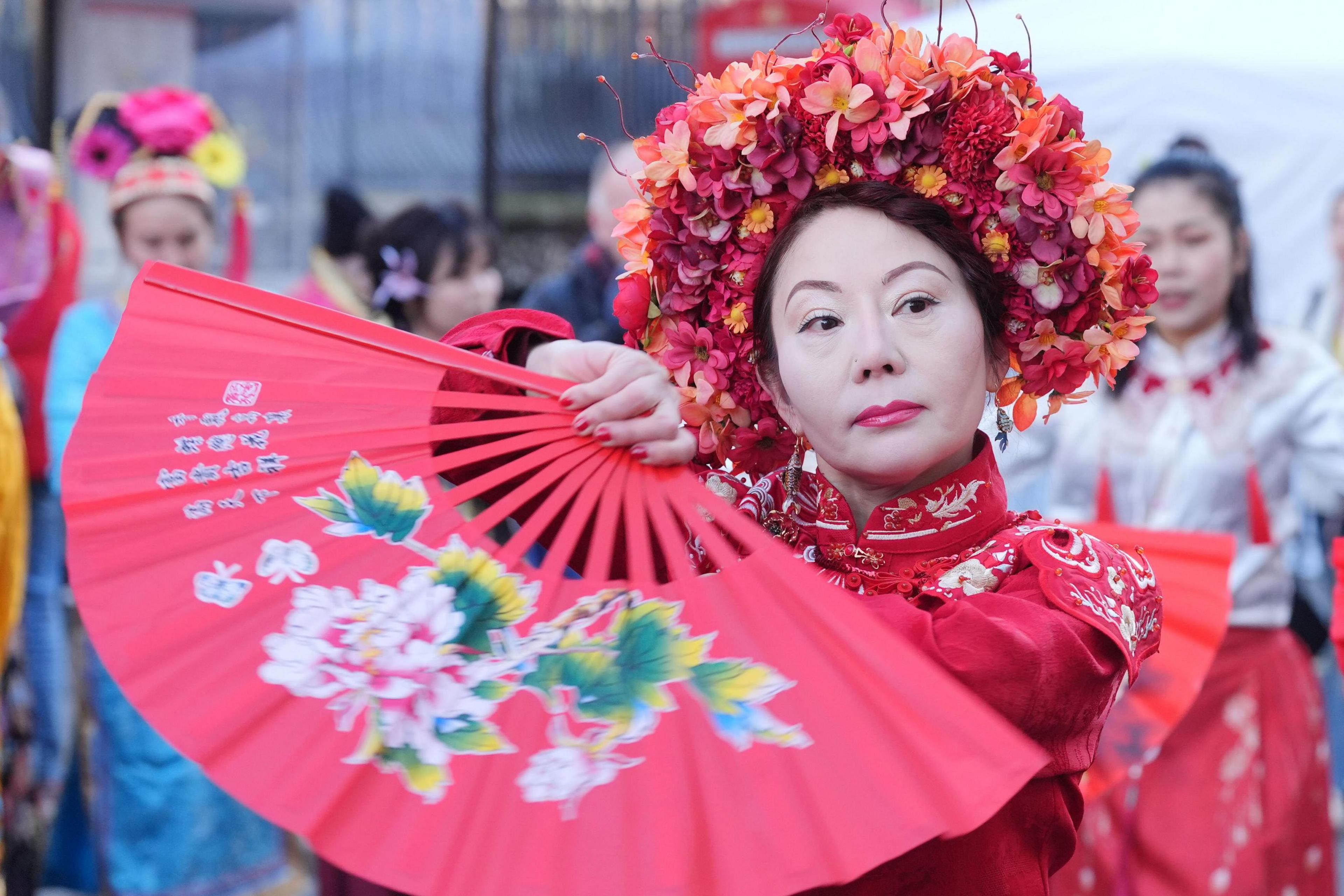 A woman dressed in a red traditional Chinese outfit holds a red folding fan decorated with floral patterns. She wears an elaborate floral headdress, and other performers in traditional attire are visible in the background.