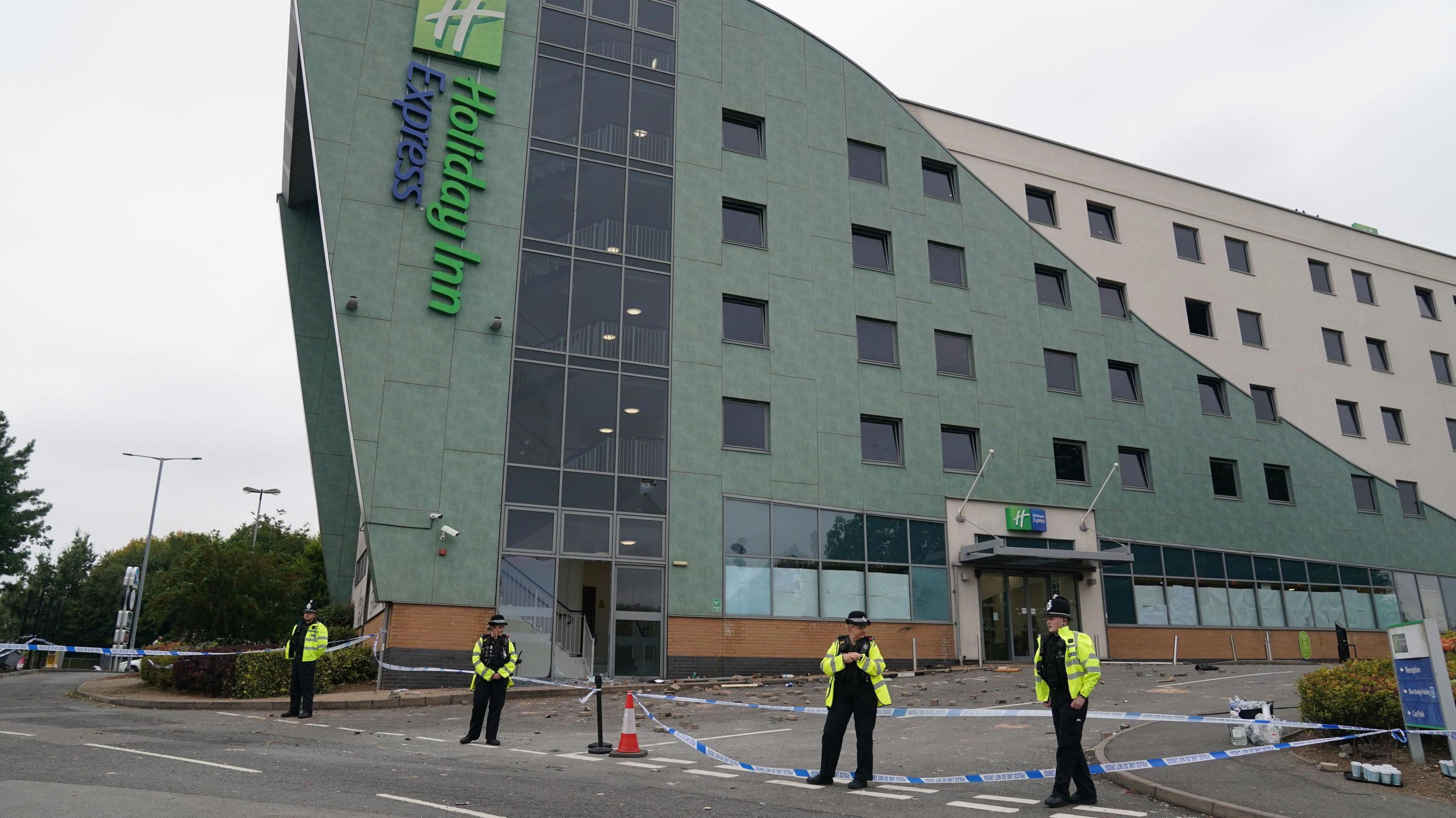 A large green and white building with a vertical "Holiday Inn Express" sign mounted on the side. Four police officers stand outside the hotel next to blue and white police tape. Behind the tape, bricks and debris is lying in the ground.