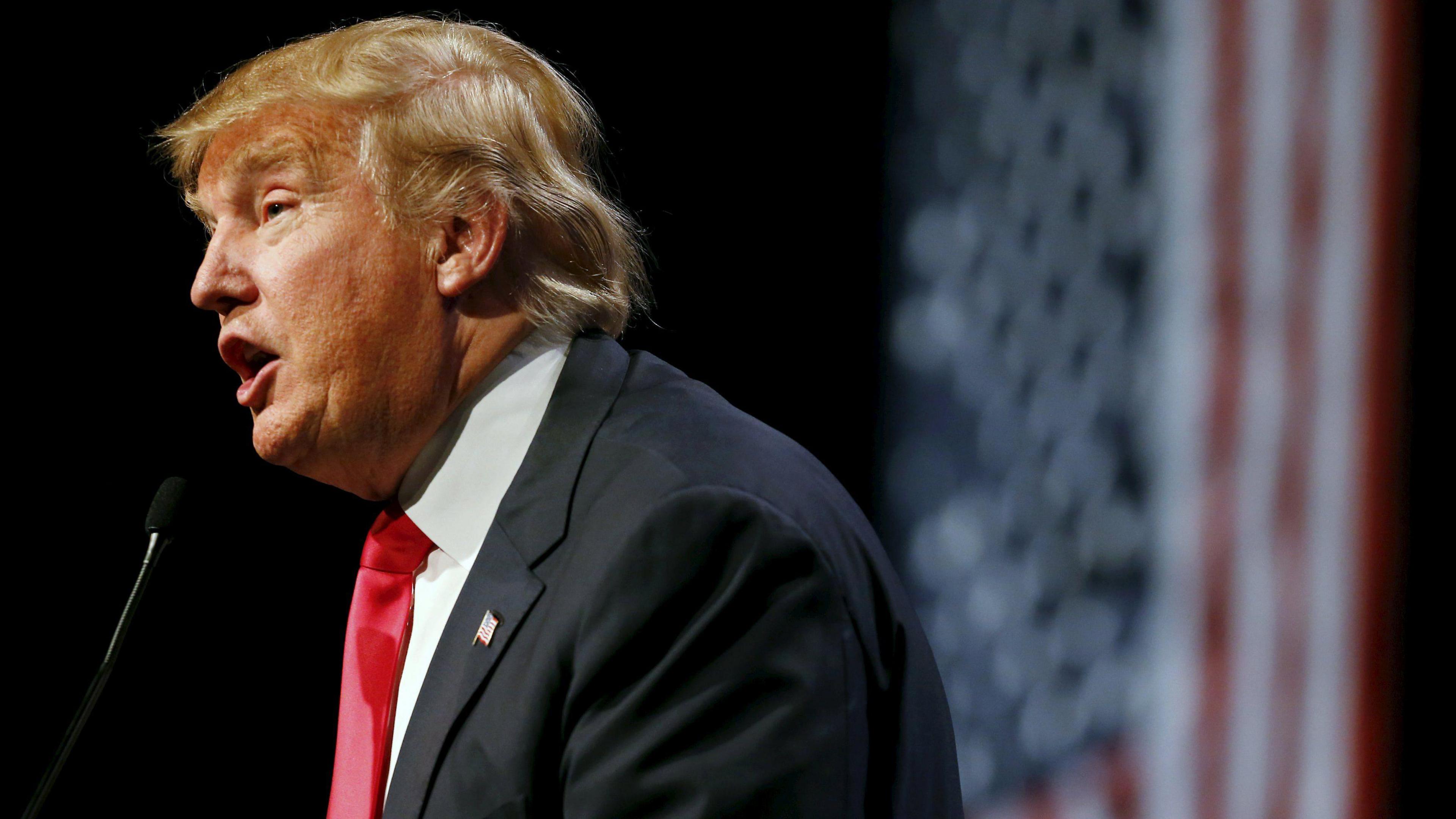 A photo of Donald trump on stage in Las Vegas in 2015. He is wearing a dark blue suit, white shirt and a red tie. In the background is an American flag
